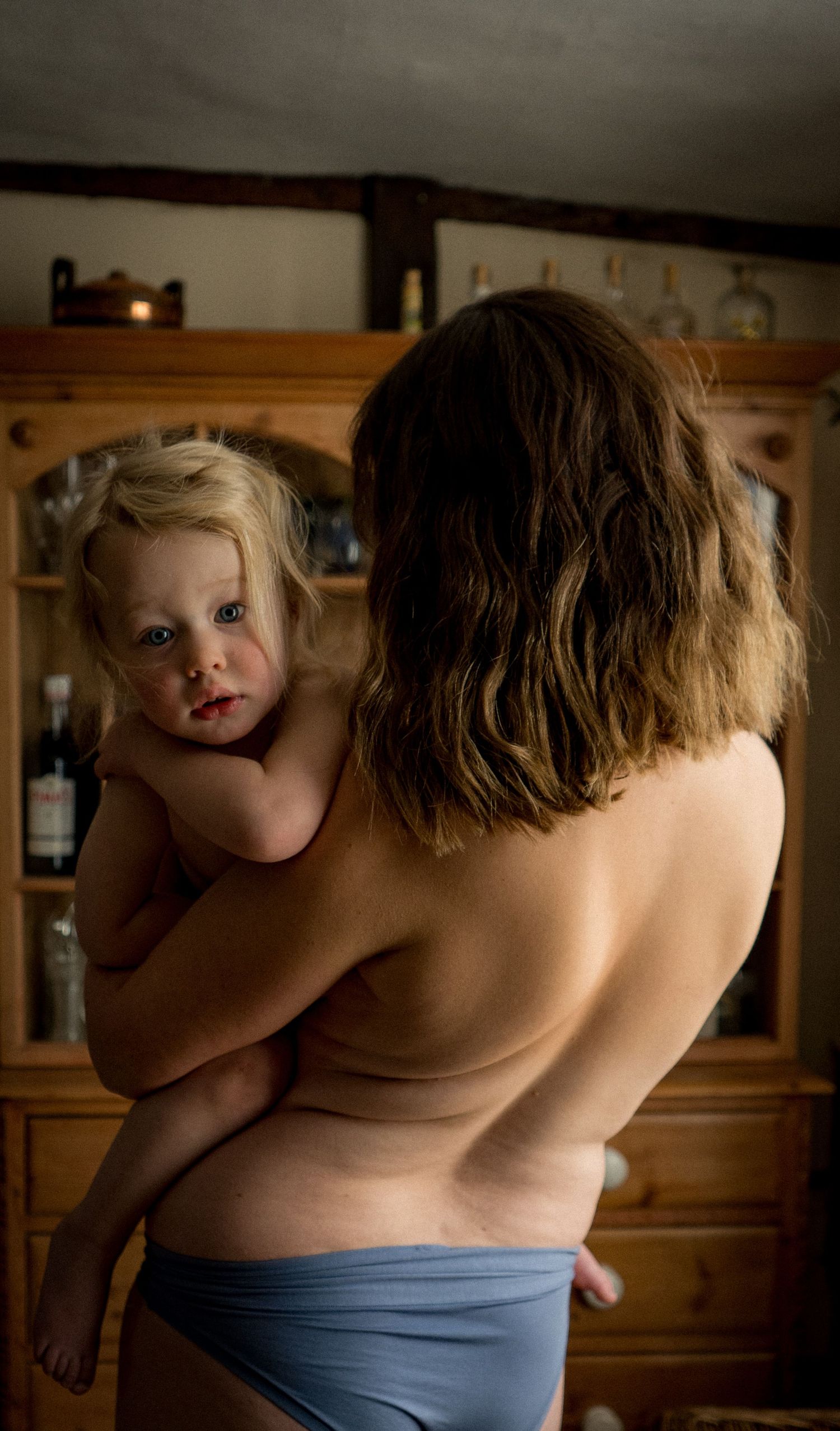 Intimate moment captured from behind showing parent in blue shorts holding young child against wooden cabinet background.