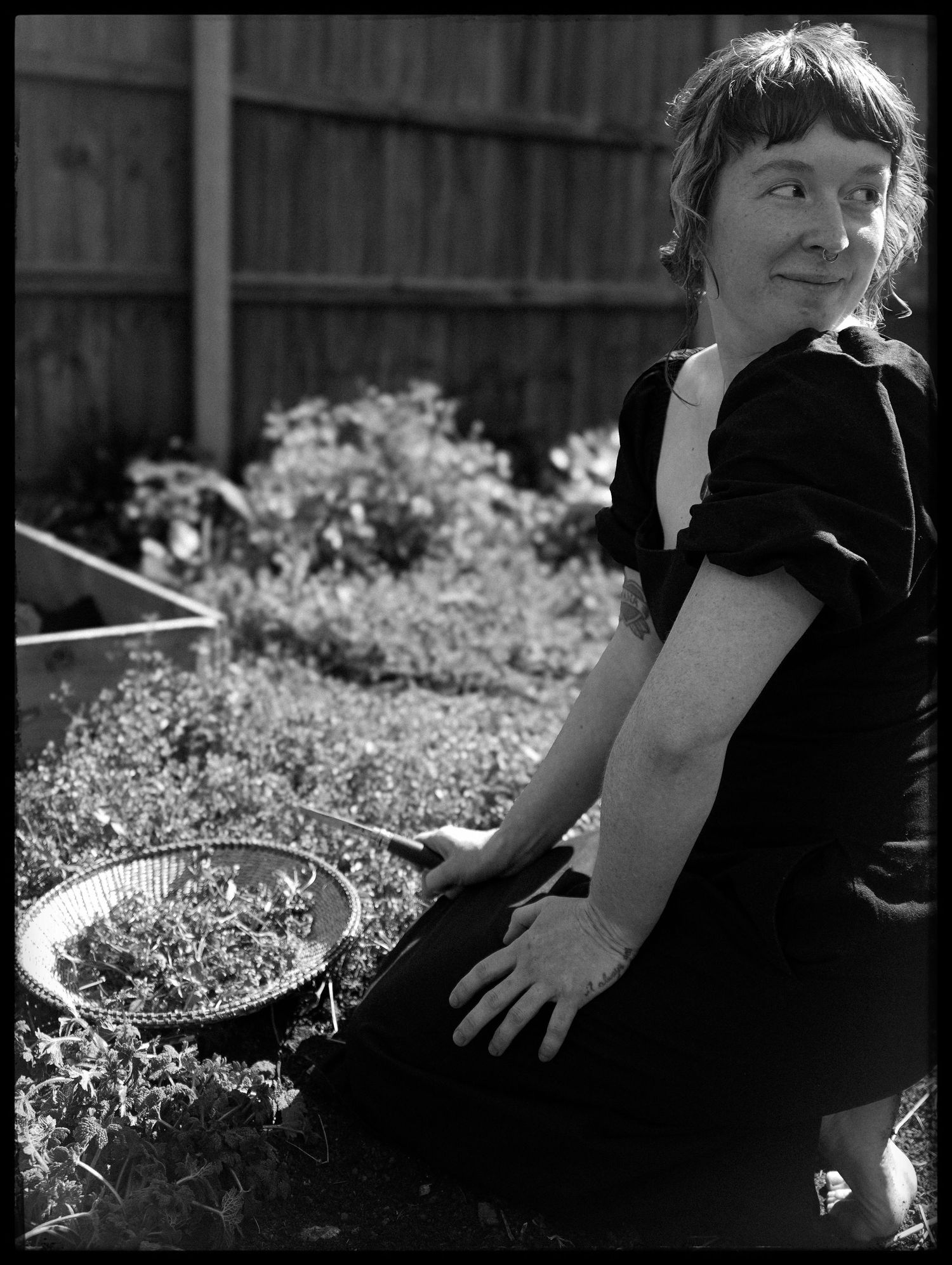 A vintage black and white photo of someone in a black dress sitting in a garden with greenery and a wooden fence behind them.