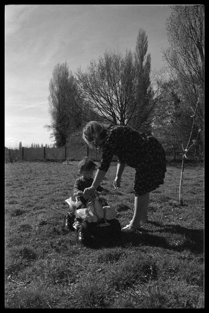 A black and white photograph shows a person bending down to interact with a small cat in a grassy field with tall trees.