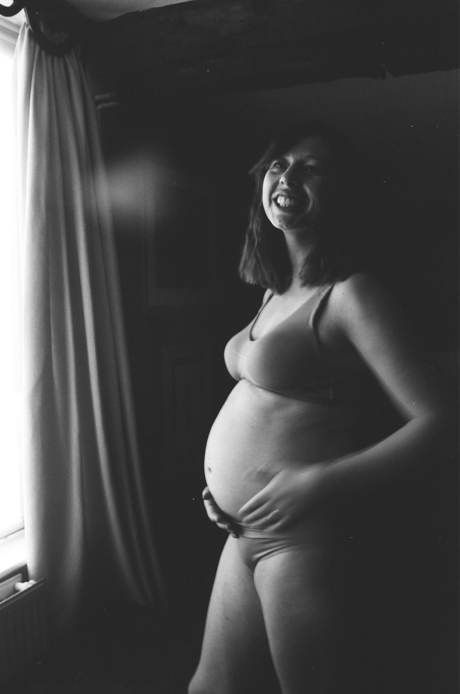Black and white photo of laughing pregnant woman in underwear standing by window with curtain.