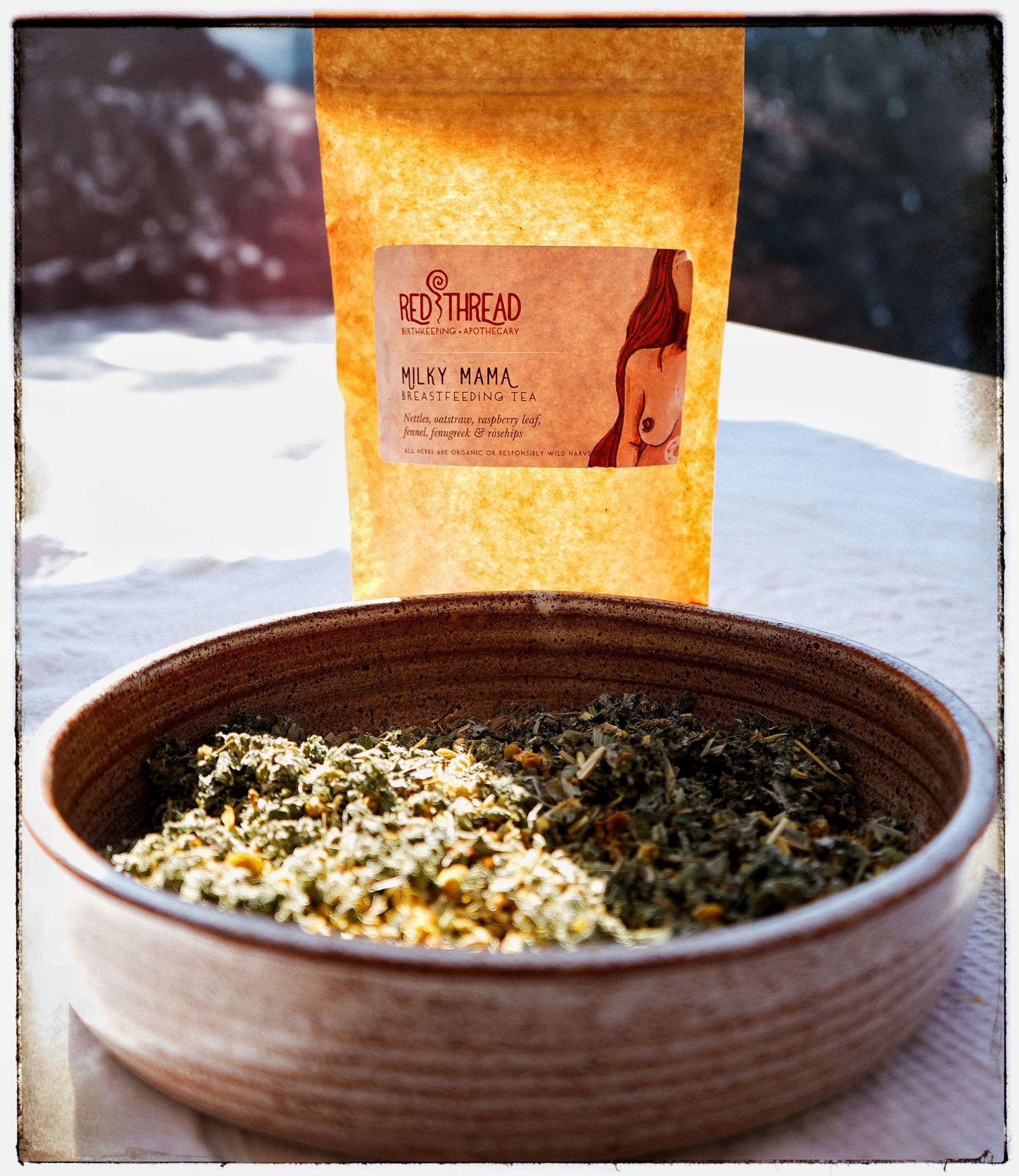 A brown ceramic bowl filled with loose herbs and tea leaves next to a golden-colored tea package with a kangaroo logo.