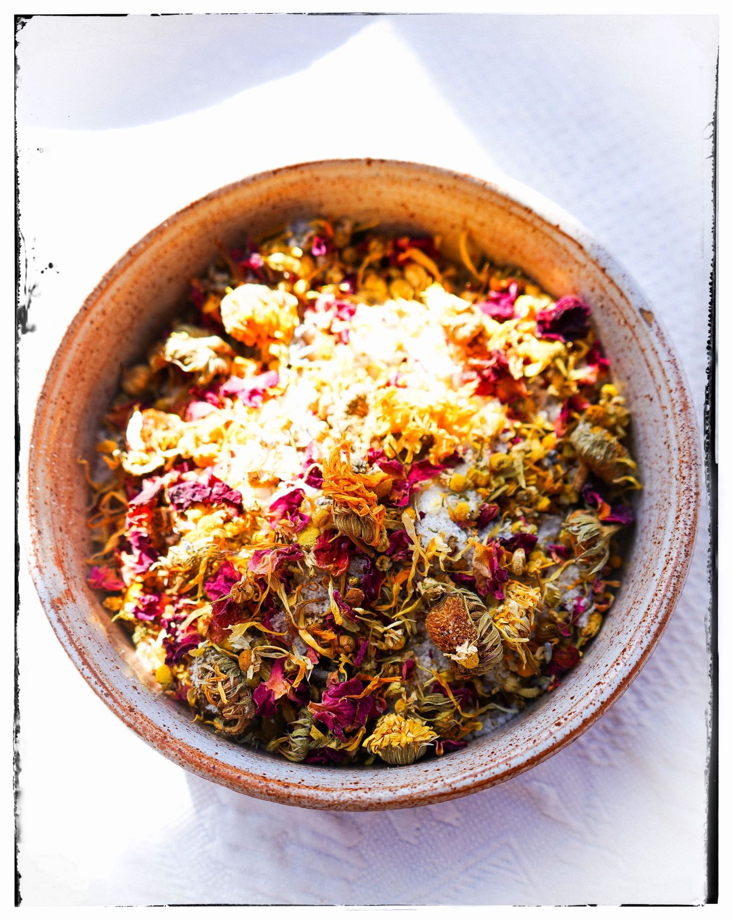 Dried herbs and flower petals arranged in a rustic ceramic bowl on a white surface.