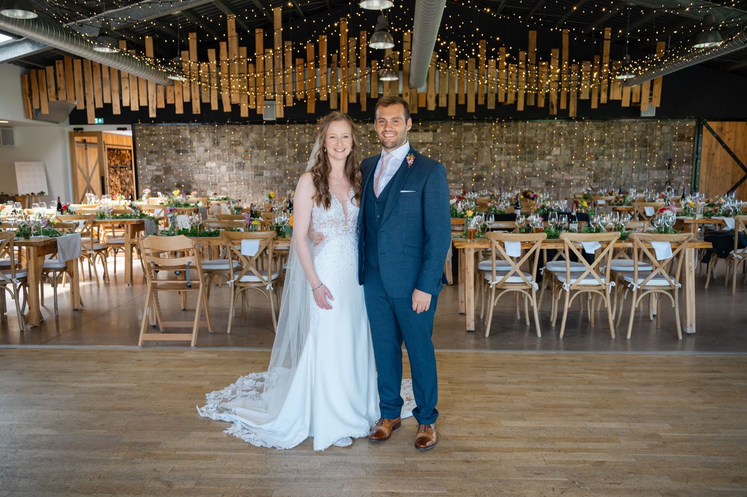 Newlyweds posing in rustic barn wedding venue with hanging lights and wooden chairs