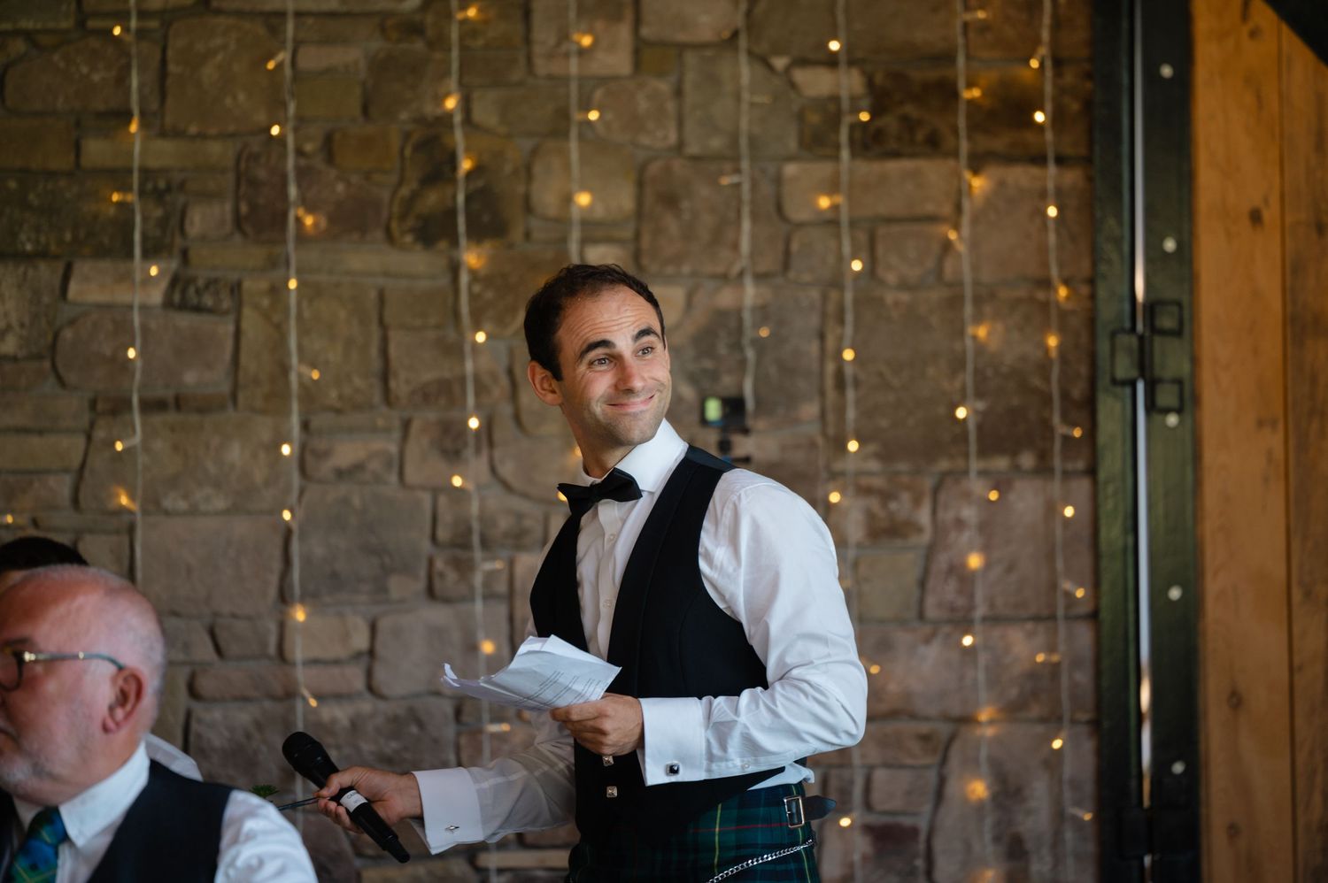 Groom standing and giving a wedding speech with soft string lights behind