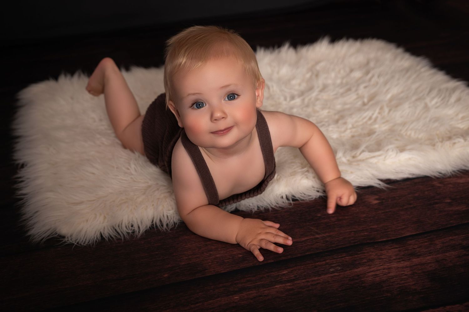 Smiling baby in a dark romper lying on a fluffy white rug, photographed in a softly lit studio with a black background.
