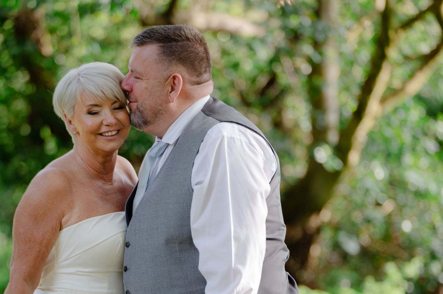 Bride and groom sharing a quiet kiss outdoors among trees, both smiling with their eyes closed in a romantic, natural moment.