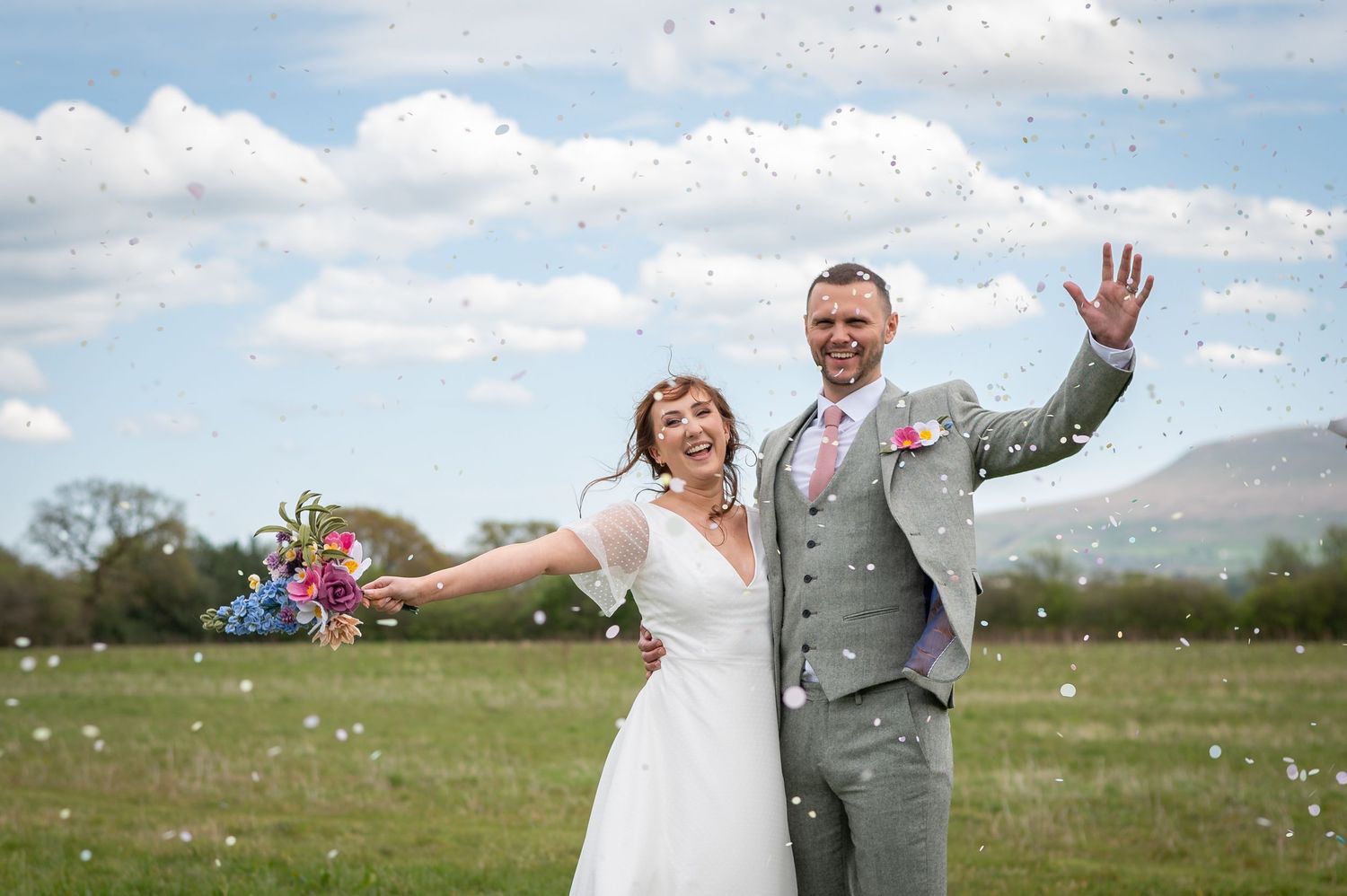 Bride and groom standing in a sunny open field, smiling and waving with arms raised, with blue skies and rolling hills in the background.