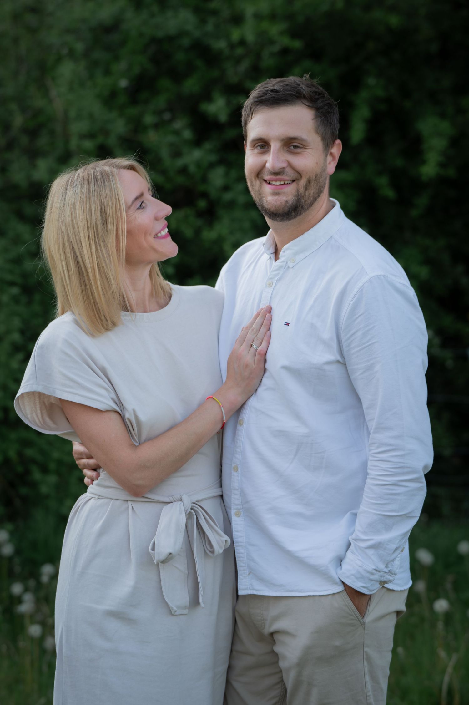 Couple standing close together outdoors, smiling warmly at the camera in coordinated white clothing, surrounded by nature.