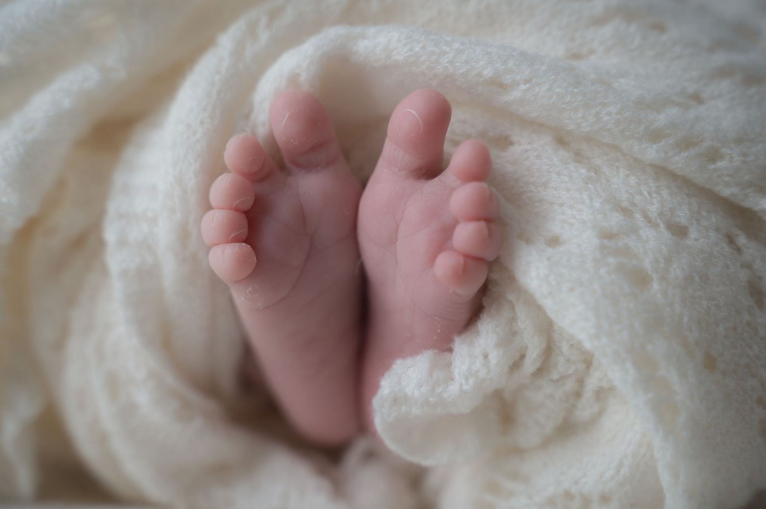 Close-up of newborn baby feet wrapped in a cream knitted blanket, showing tiny toes with soft natural light.