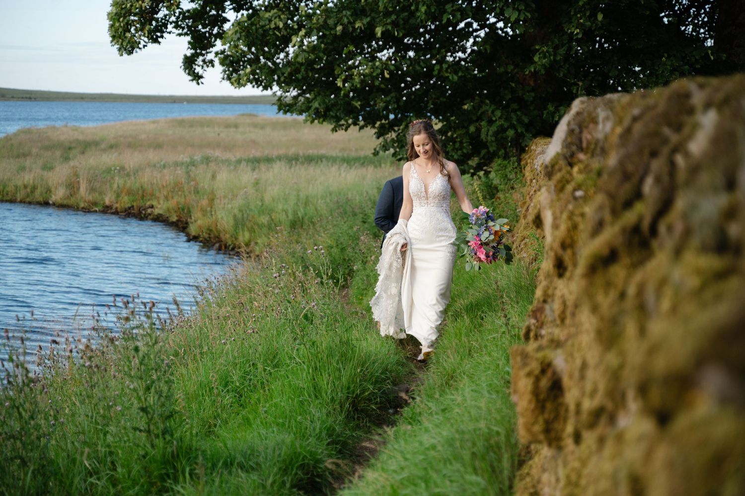 Bride walking alone on countryside path beside river and stone wall