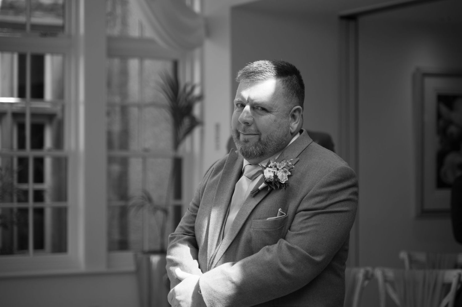 Black and white portrait of the groom standing indoors with arms crossed, wearing a boutonnière and smiling slightly.