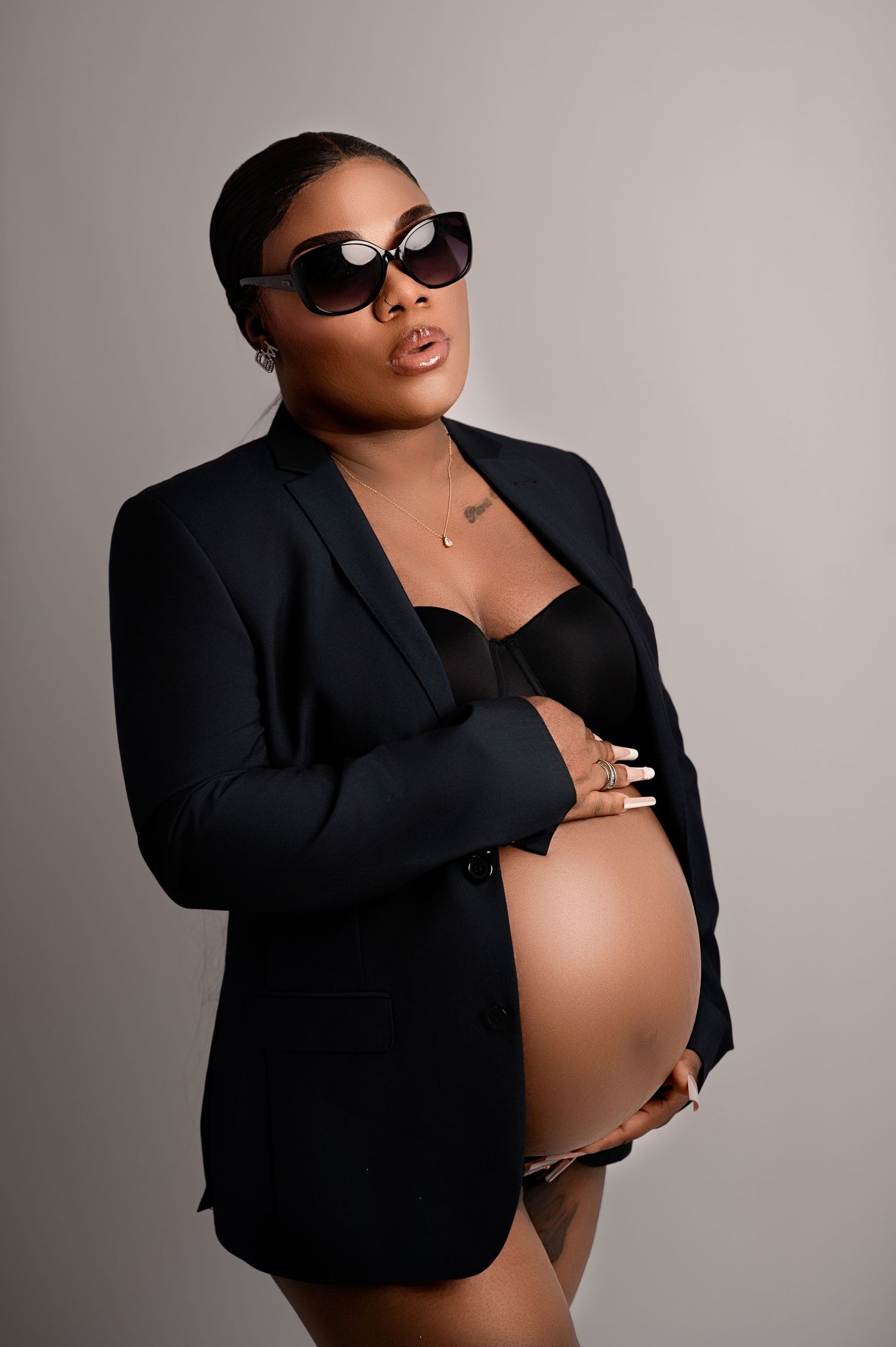 Pregnant woman in a black blazer and sunglasses, posing boldly with hand on chest and bare belly showing.