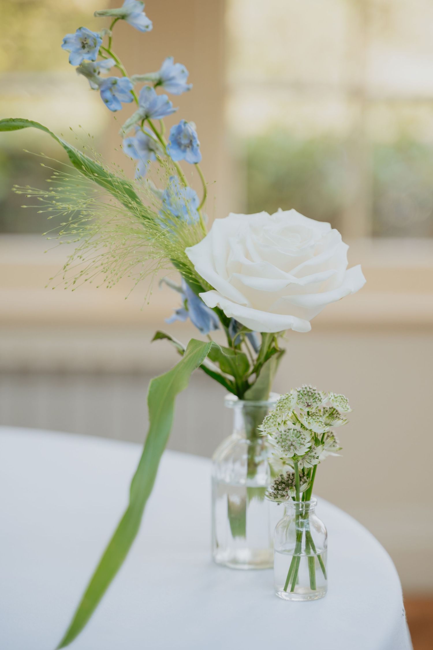 Minimalist wedding table decor featuring small glass vases with single stem white and blue flowers on a crisp white tablecloth.