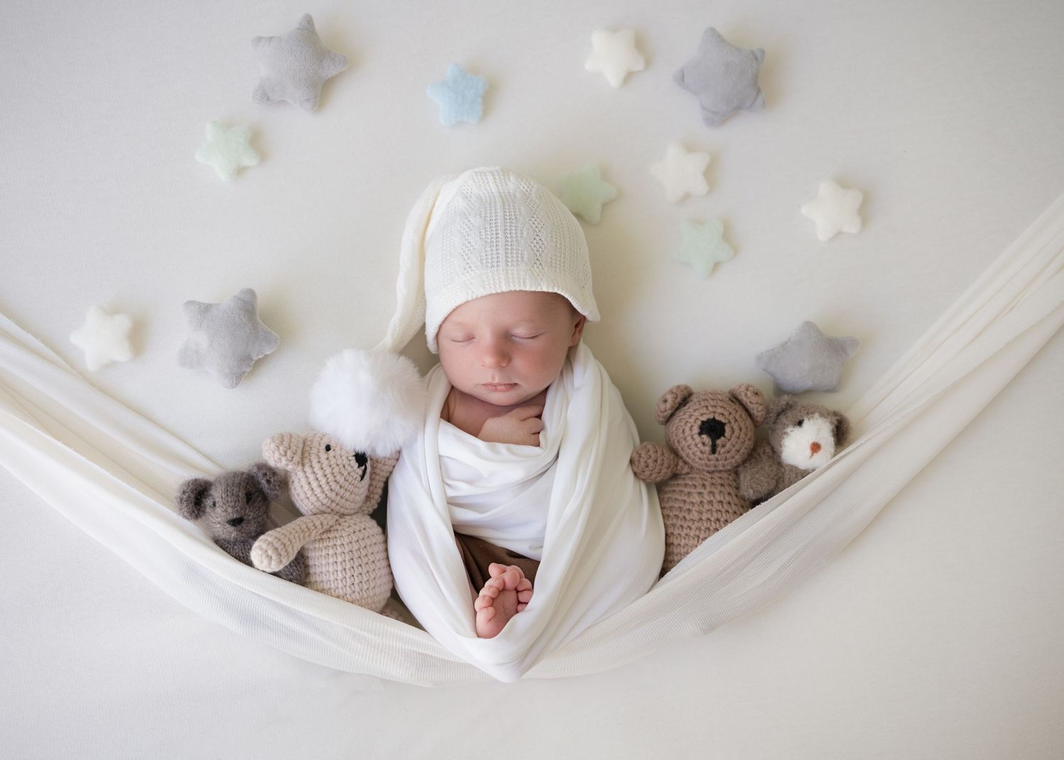 Newborn baby wrapped in soft white fabric, resting in a hammock with teddy bears, surrounded by soft star decorations in a dreamy studio set