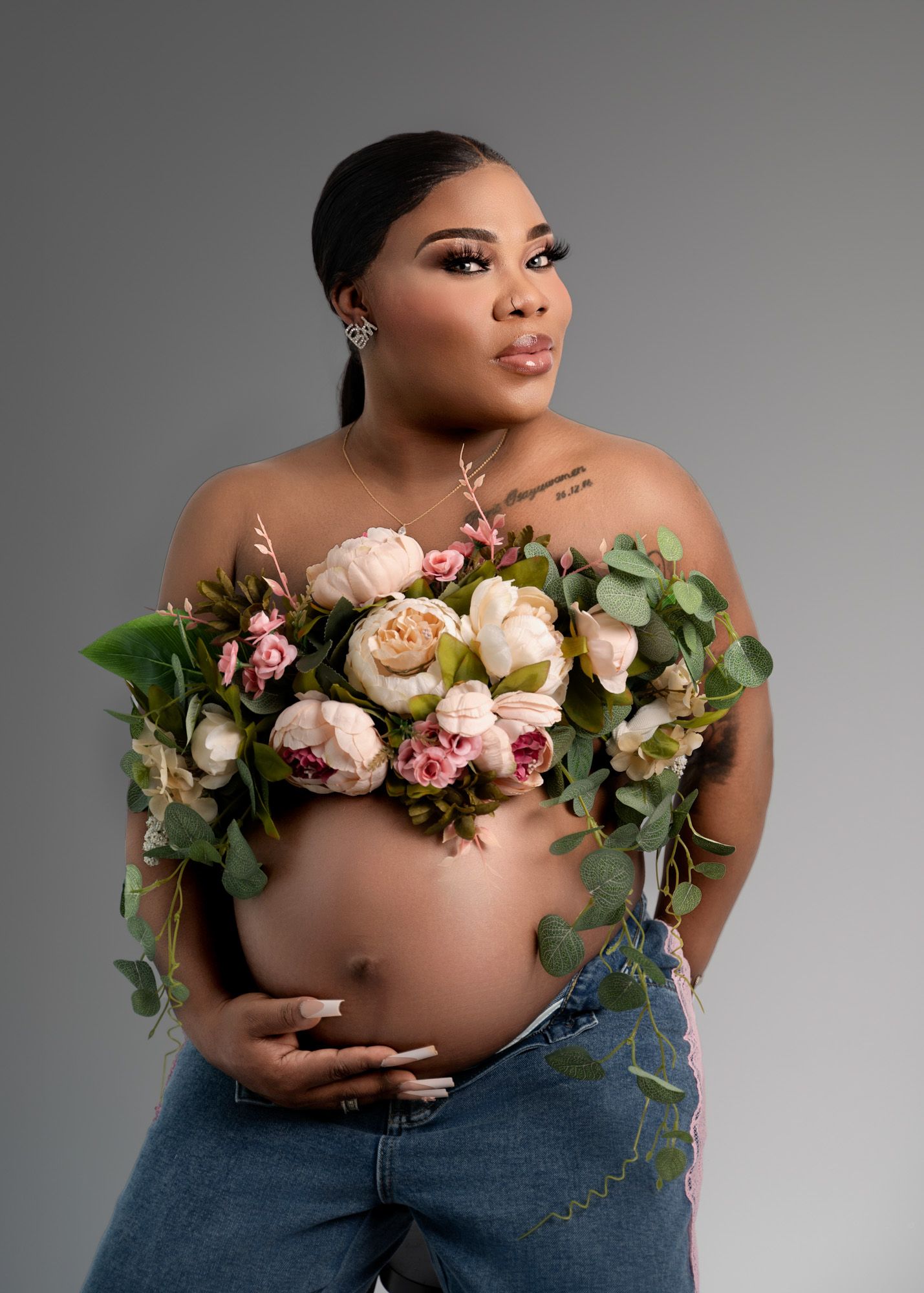 Pregnant woman in jeans with floral top wrapped around her chest and belly, looking off-camera against a gray background.