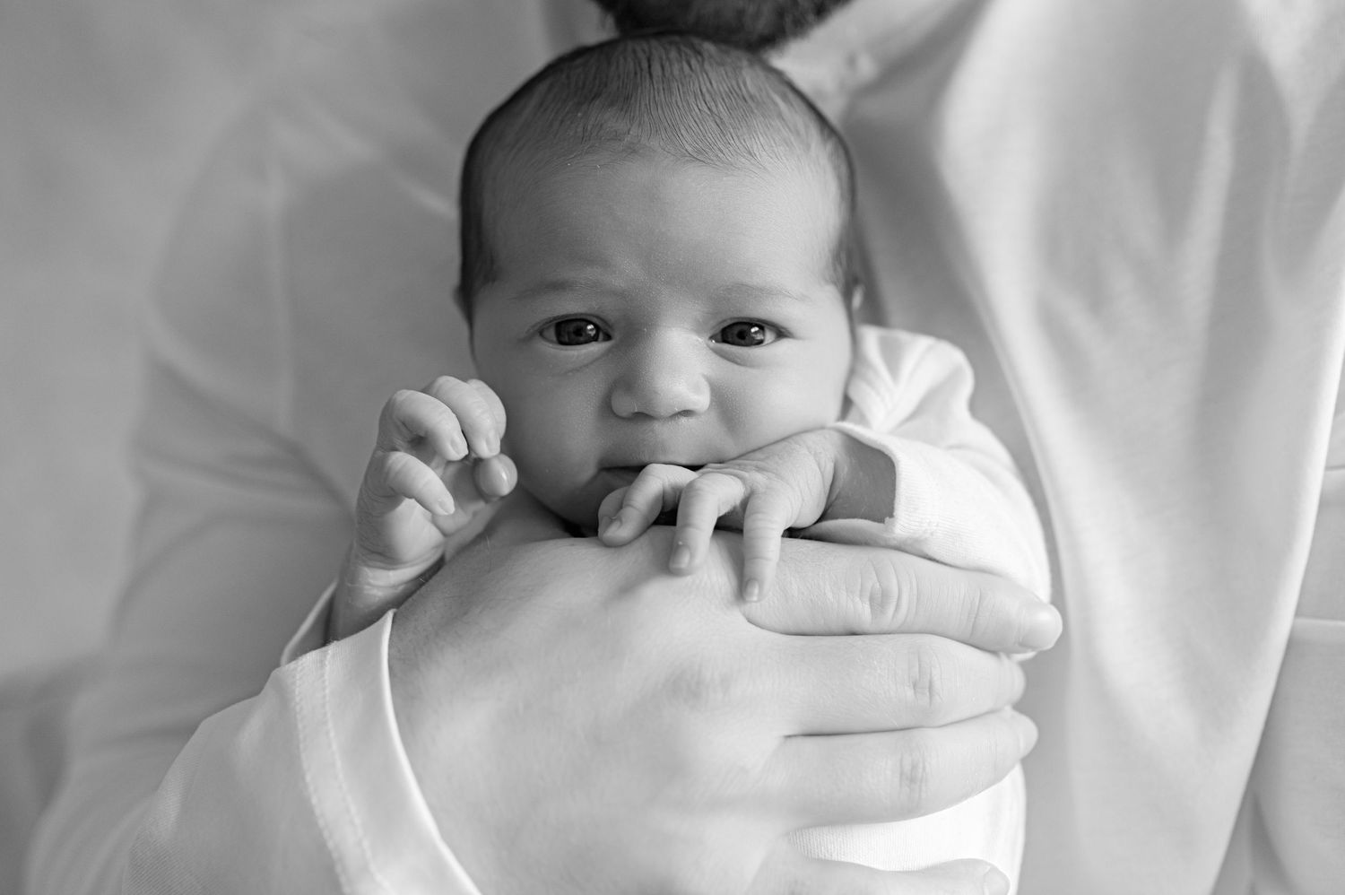 Black and white photo of a wide-eyed baby lying on a bed, holding a soft toy and looking directly at the camera.