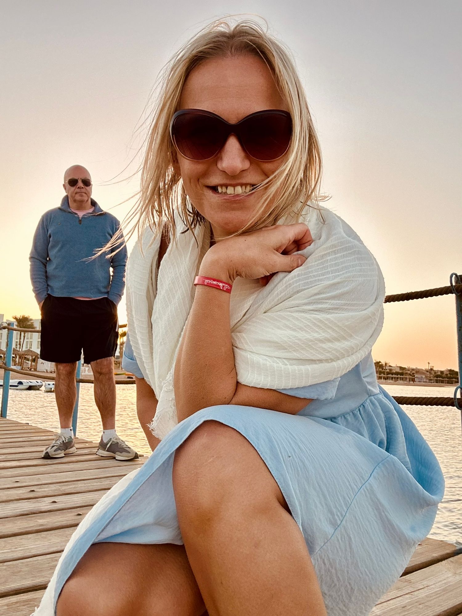 Woman sitting on a wooden boardwalk at sunset, smiling and wearing sunglasses and a light blue dress, with a man walking in the background.