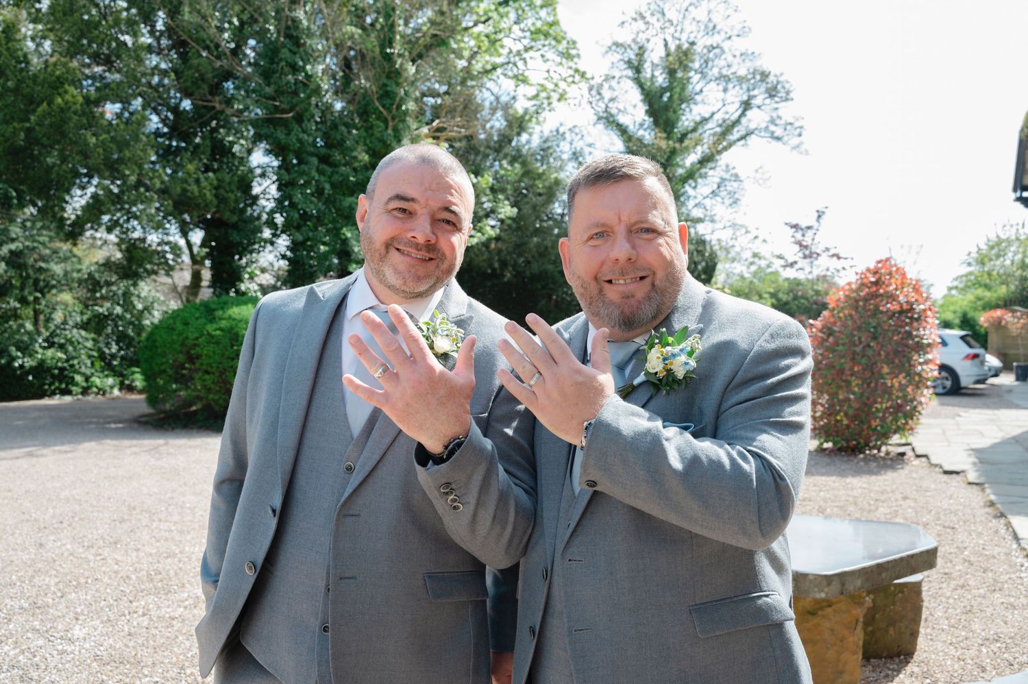 Groom and best man or guest smiling and proudly showing off their rings outdoors in natural light, dressed in grey wedding suits.