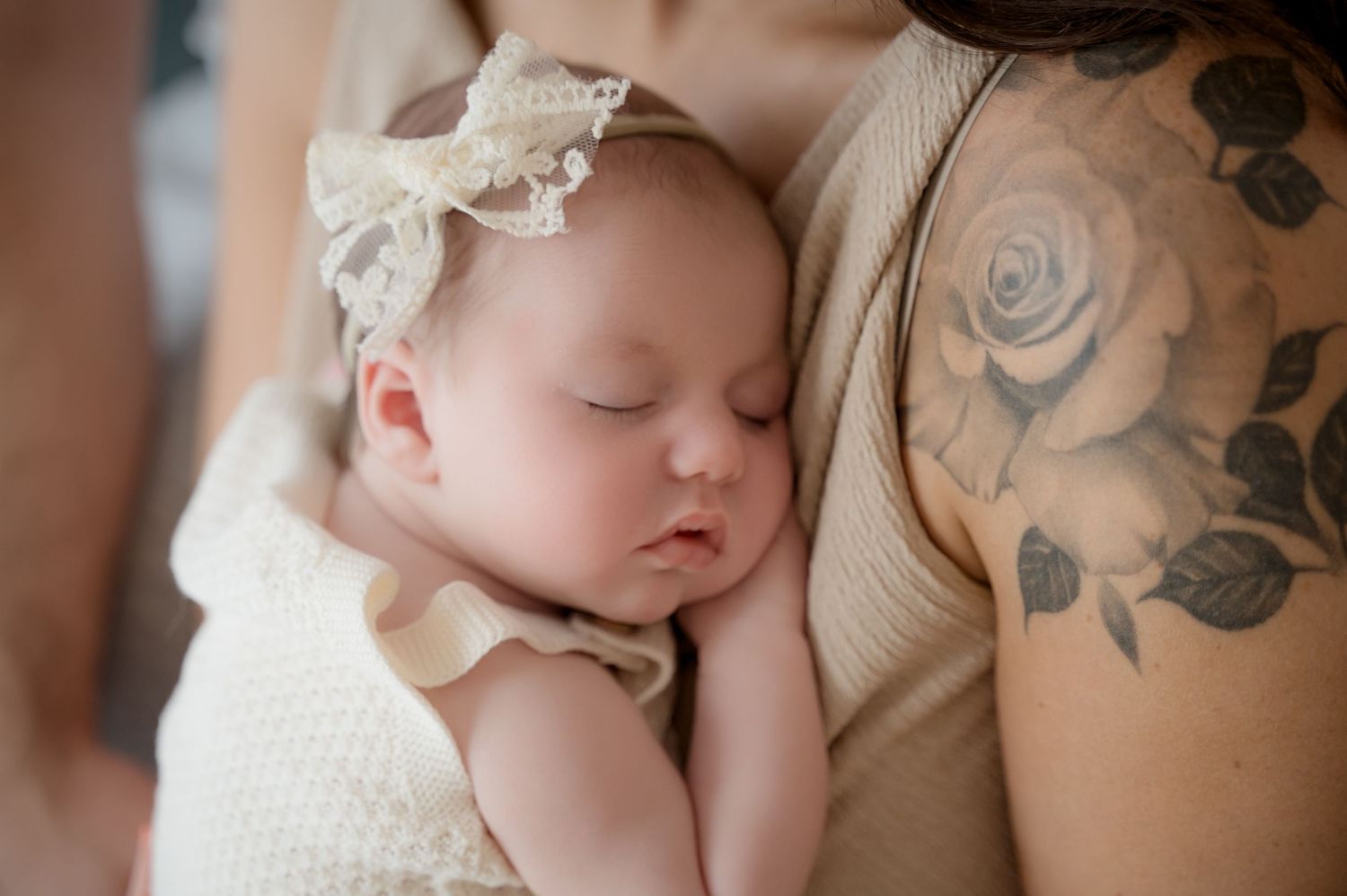 Newborn baby girl sleeping peacefully against her tattooed mother’s arm, wearing a cream bow headband and ruffled outfit