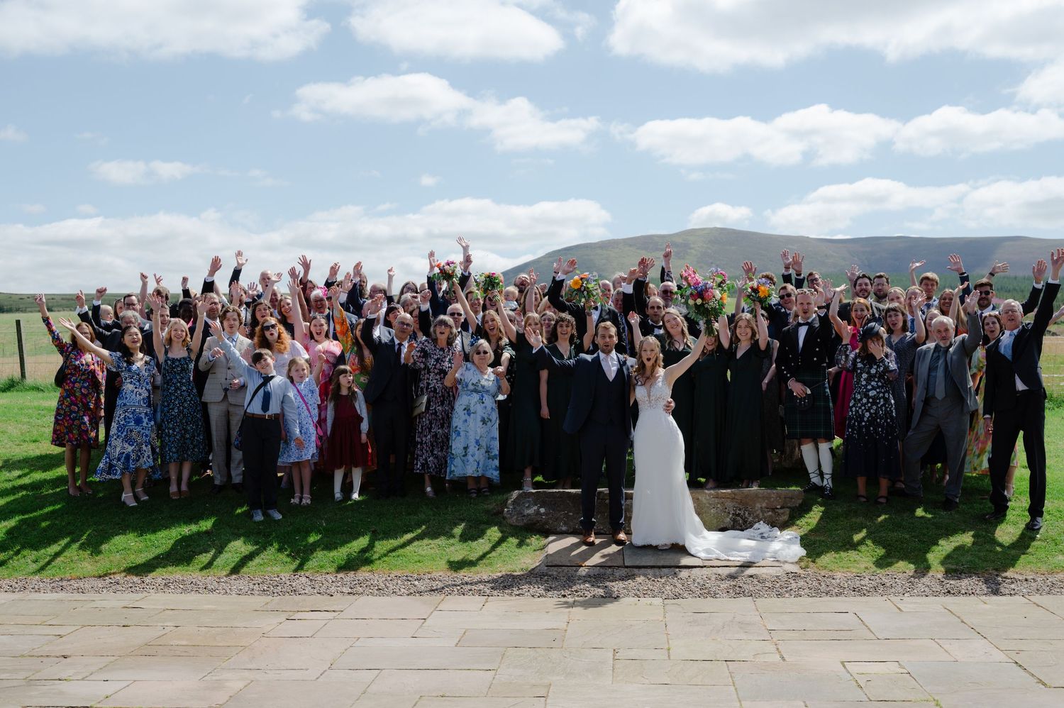 Full wedding guest group photo with bride and groom in the centre, all cheering