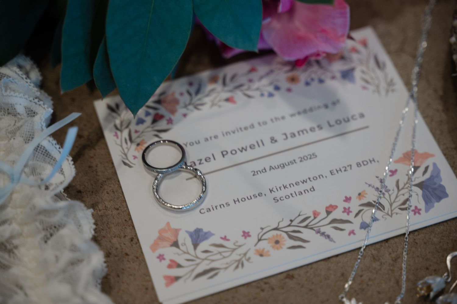 Flat lay of two wedding rings placed on a floral illustrated invitation alongside lace fabric and a velvet ring box