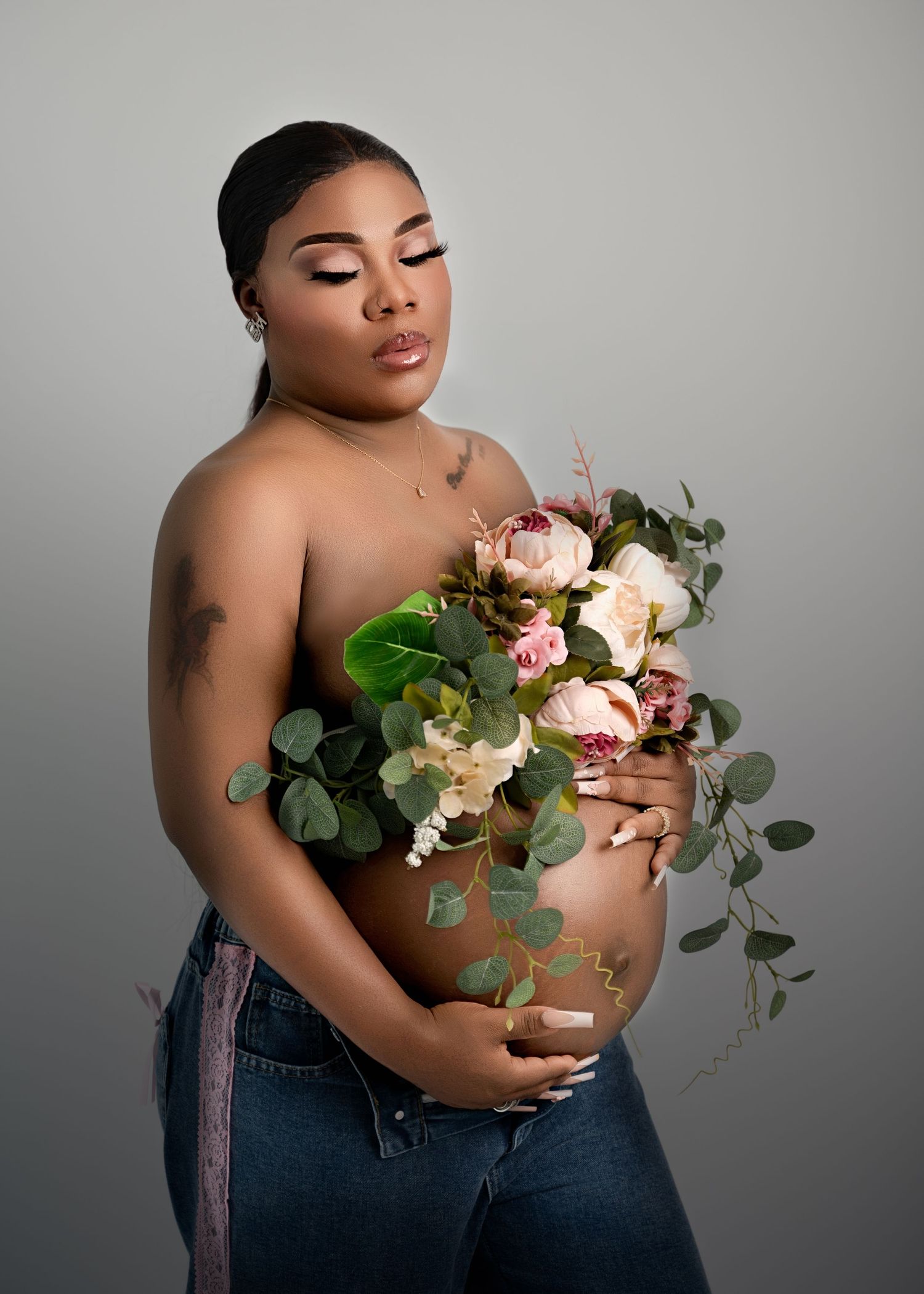 Pregnant woman holding a large floral bouquet in front of her bare belly, standing tall against a soft gray background.