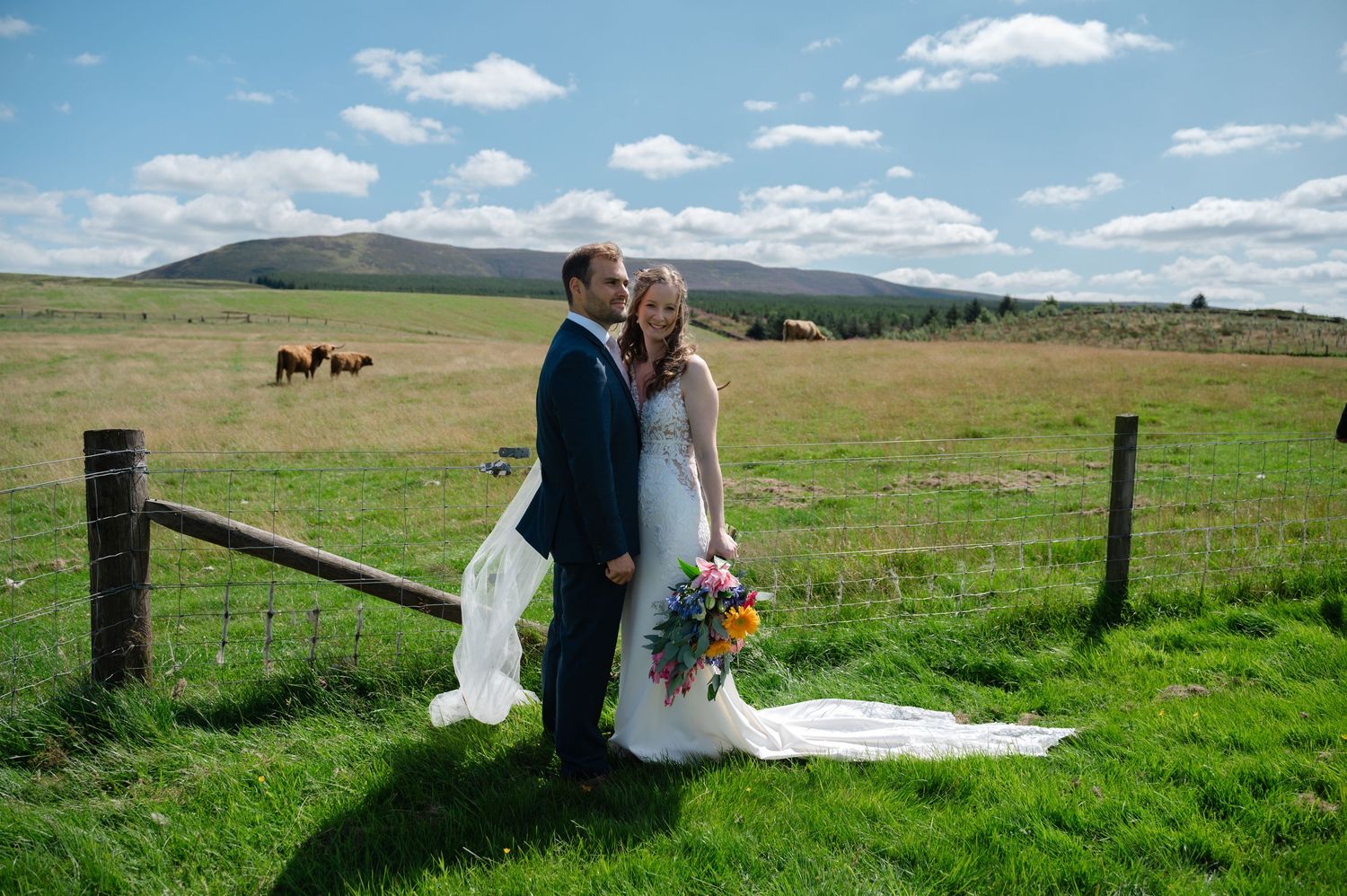 Bride and groom embracing in open field with rolling hills and blue sky
