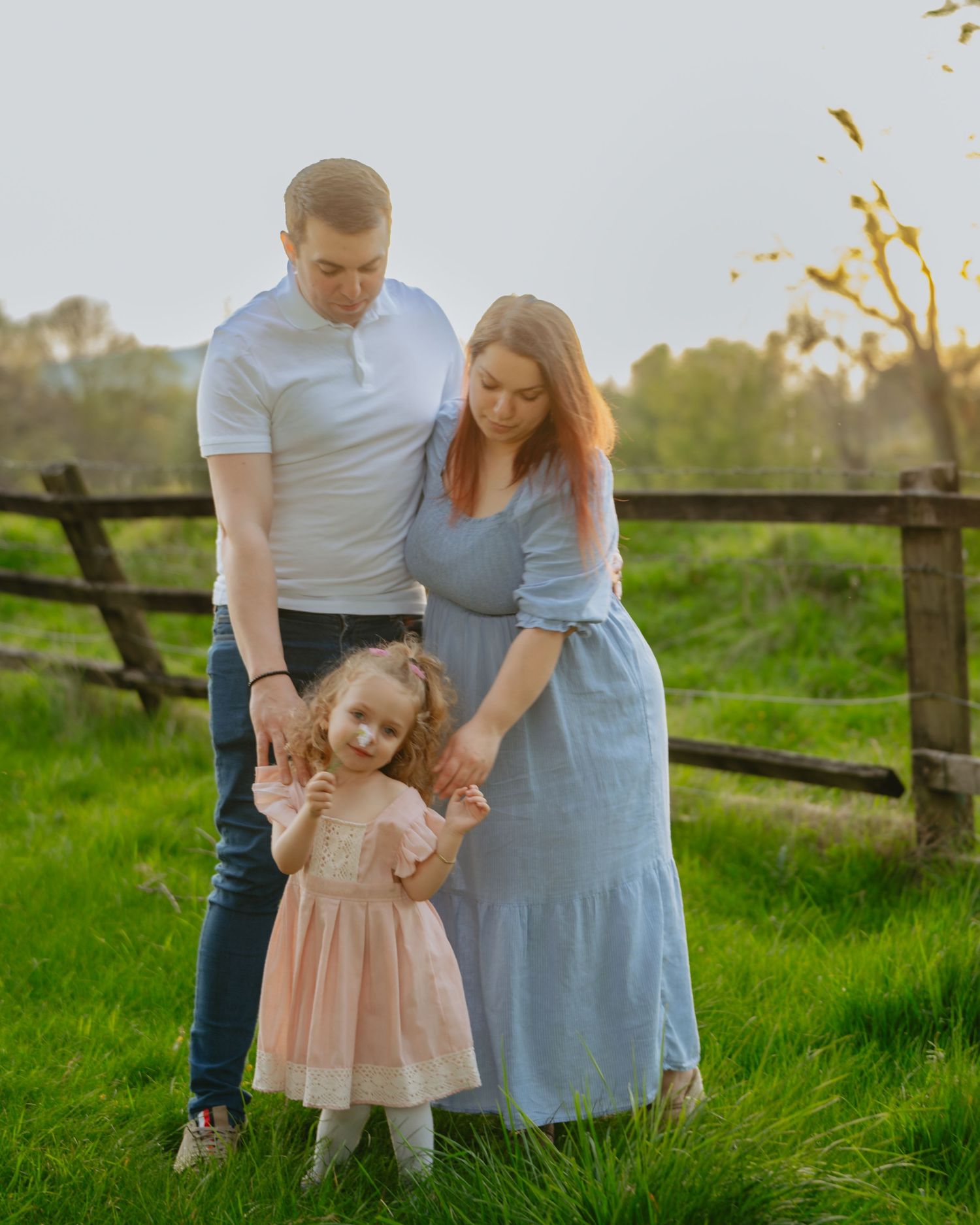 Family of three standing in a lush green field, smiling together under spring blossoms with warm golden-hour light.