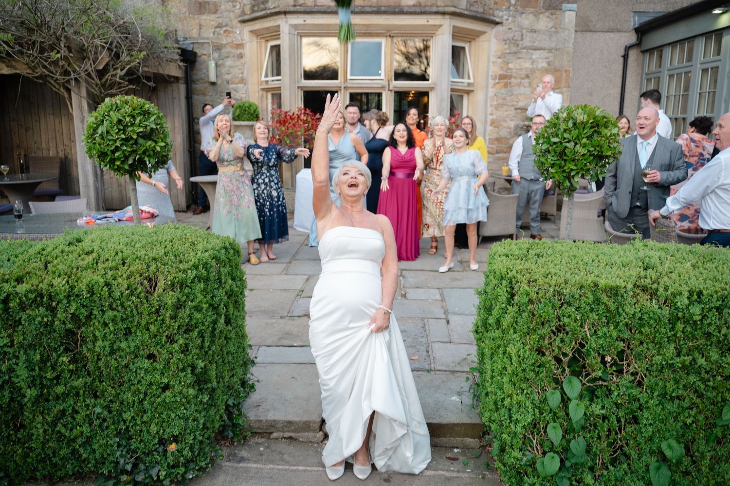 Bride throwing flowers toward a group of guests outside a wedding venue, smiling as friends and family look on.