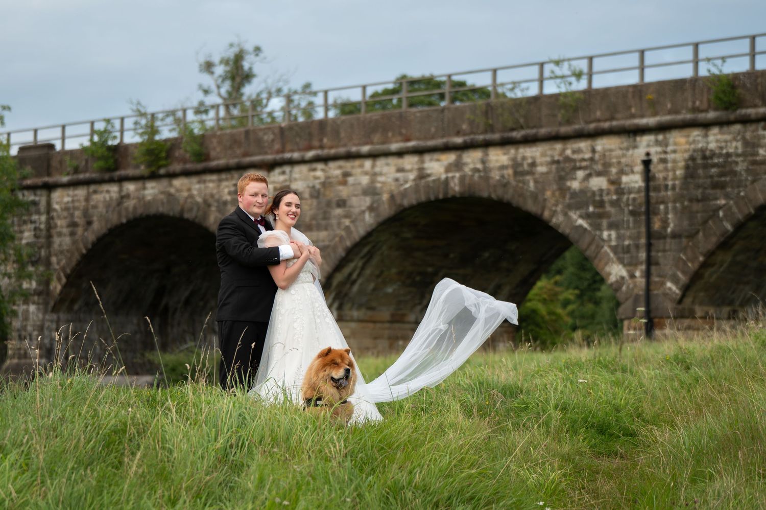 Bride and groom walking through meadow with their dog leading the way