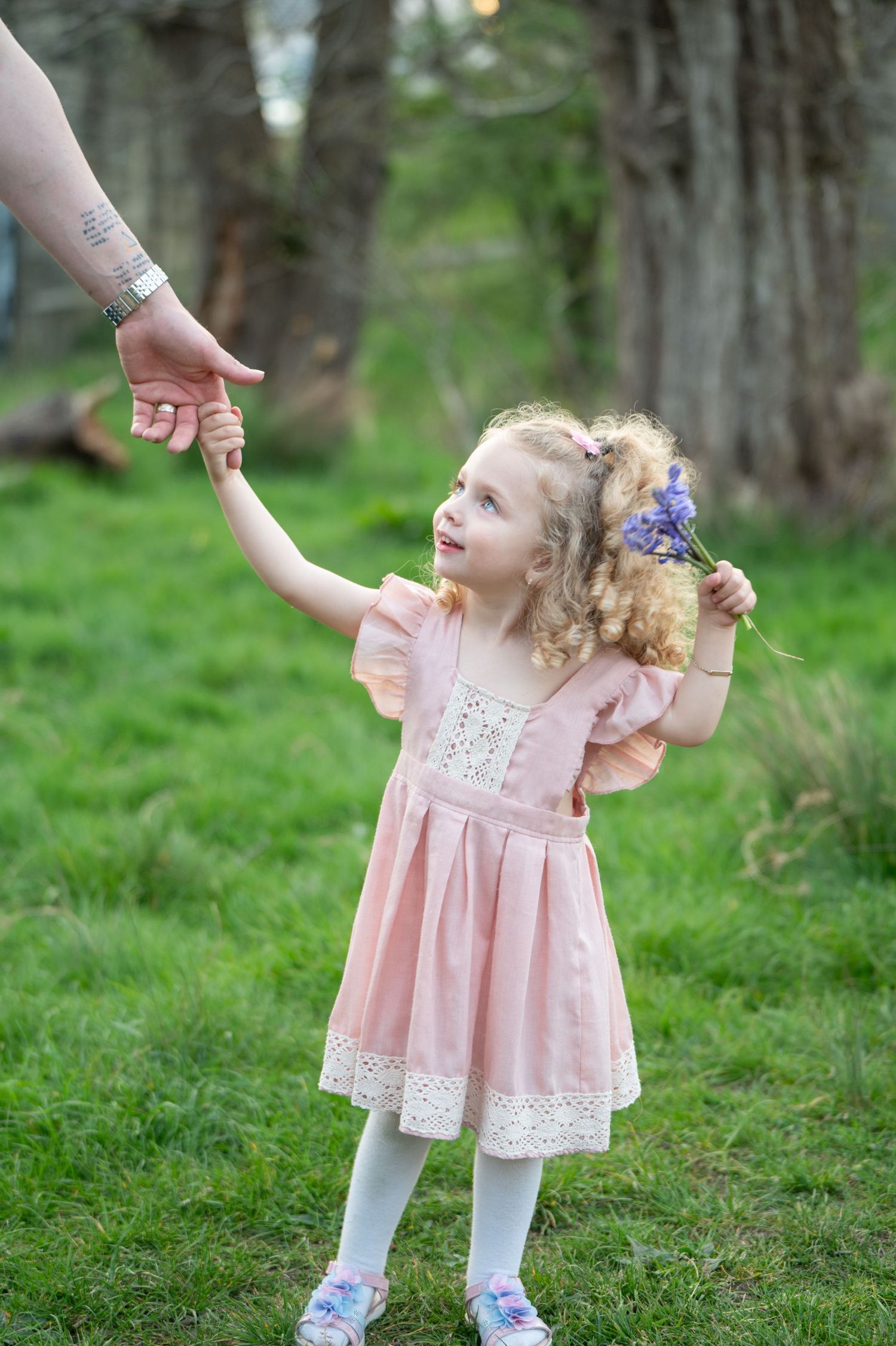 Young girl in a pink dress holding a parent's hand while walking through a grassy field, looking up playfully with a flower headband.