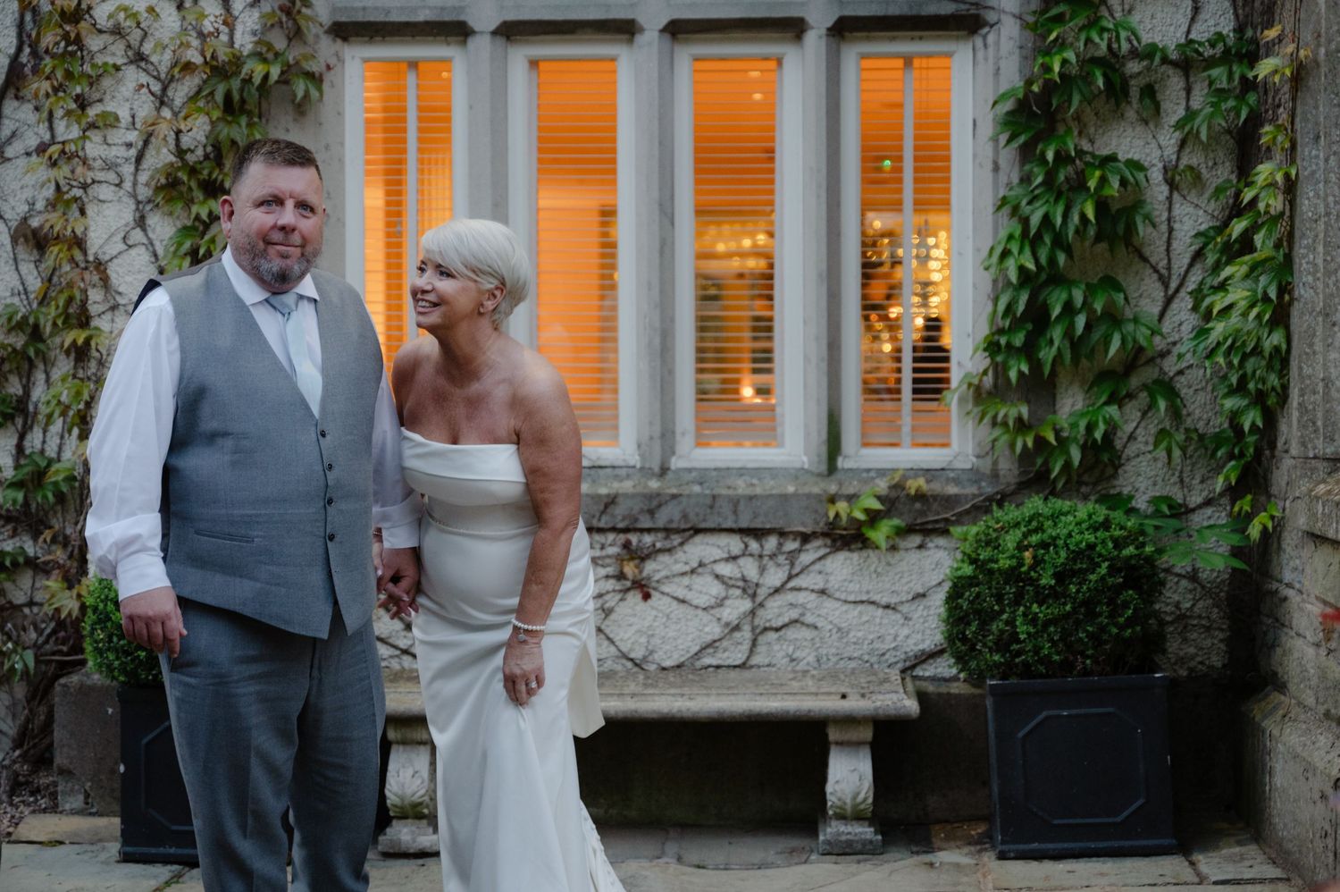 Bride and groom posing together outside a historic building with glowing windows, both dressed in formal wedding outfits.
