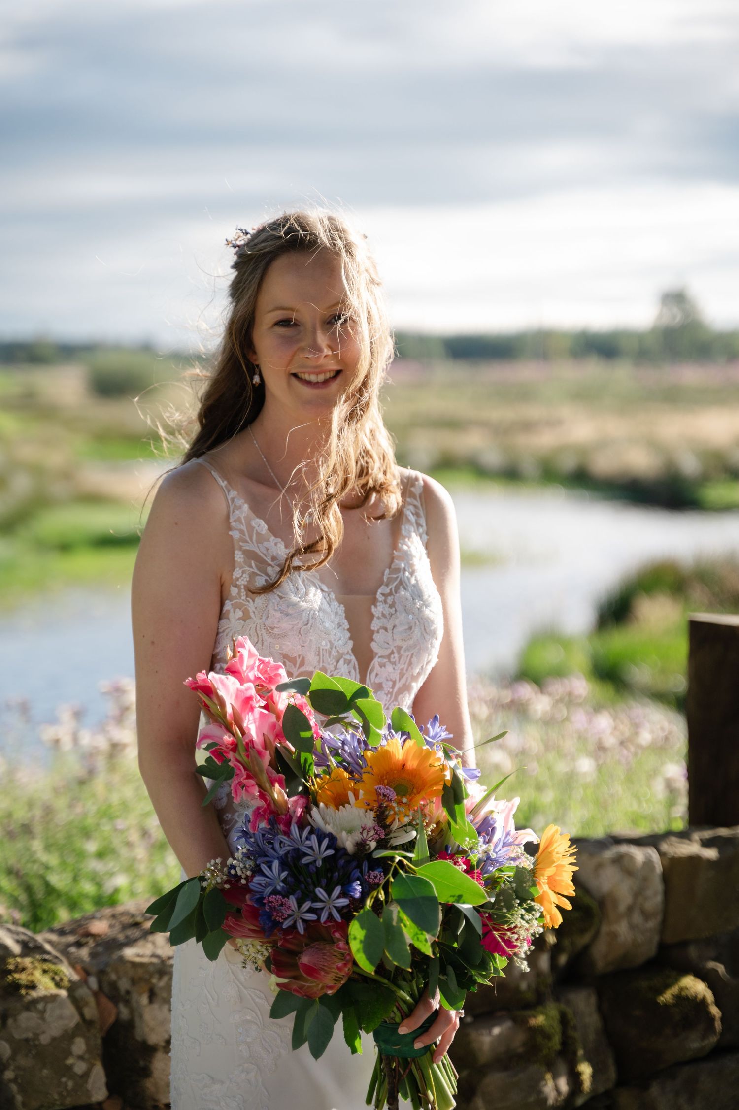 Bride standing outdoors with colourful bouquet and scenic countryside backdrop