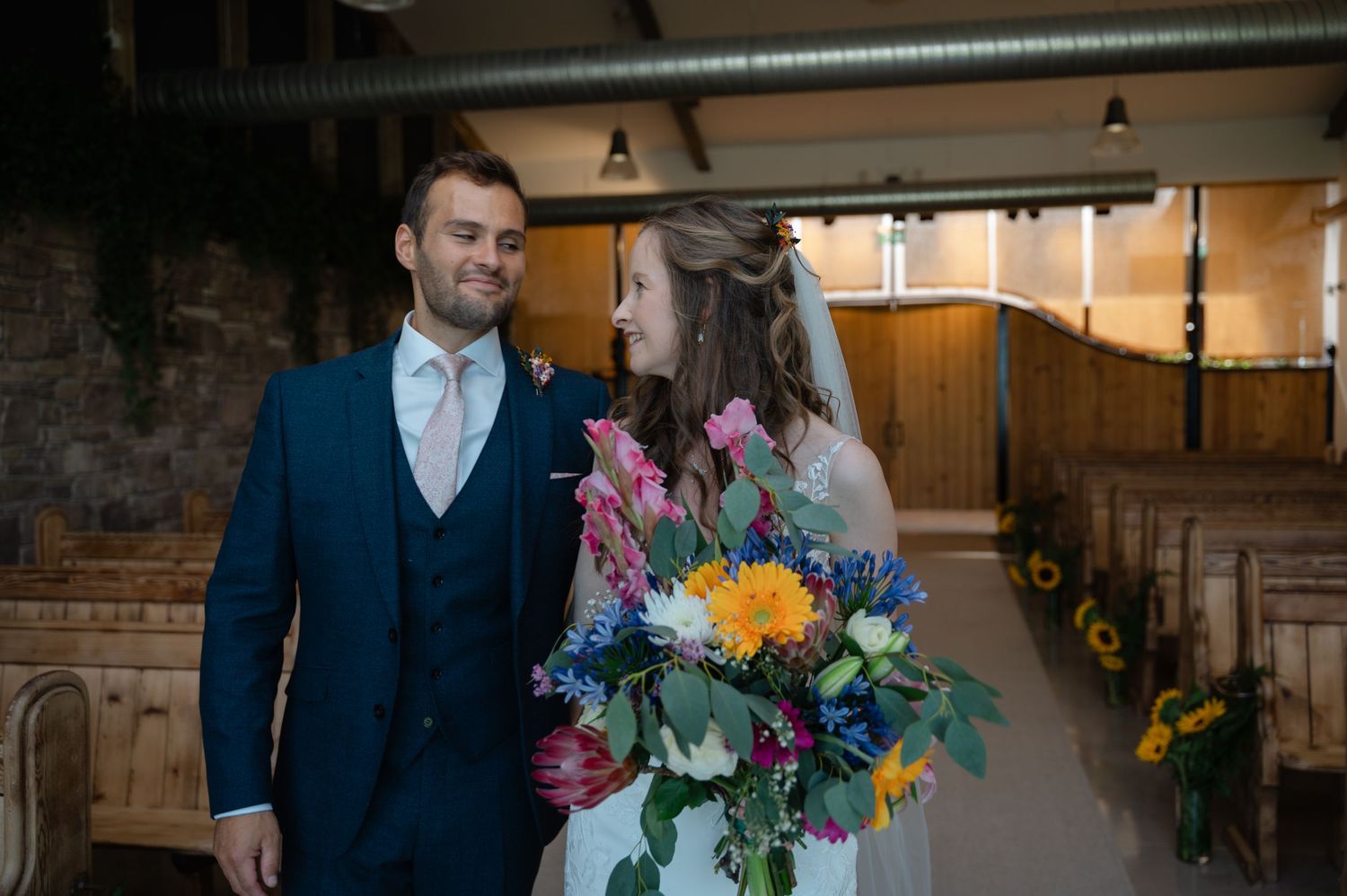Newlyweds walking down barn aisle holding vibrant floral bouquet