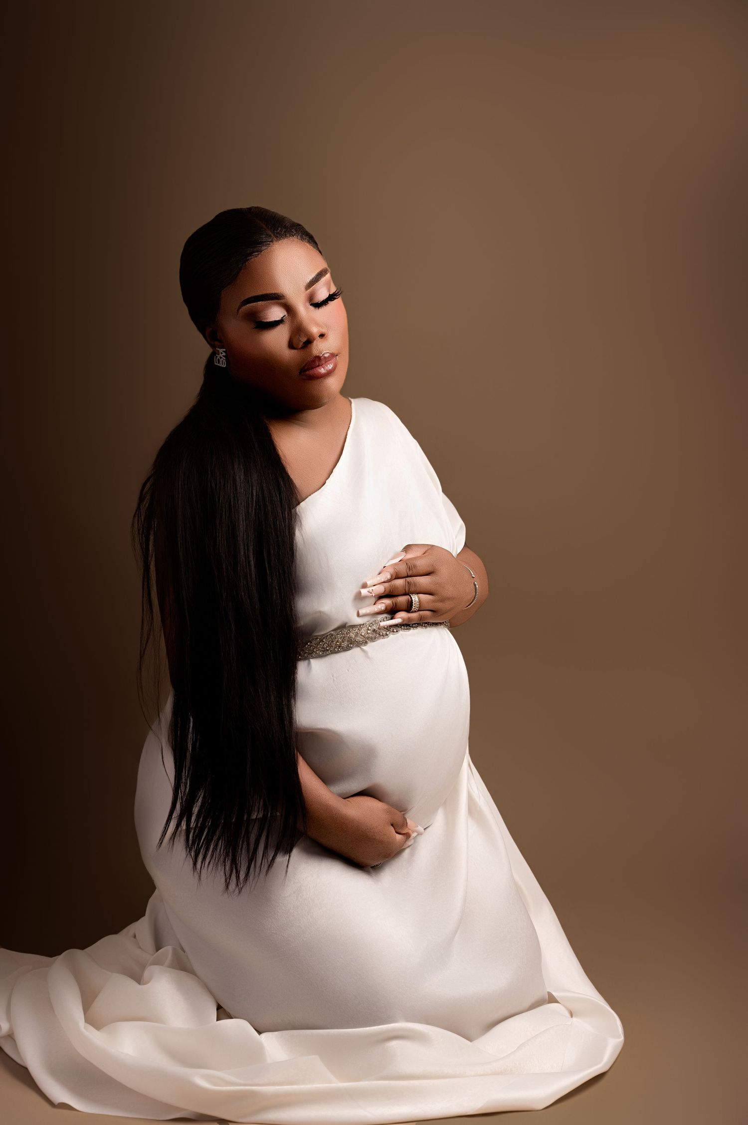 Pregnant woman kneeling in a white gown with a black shawl, smiling softly against a neutral beige backdrop.