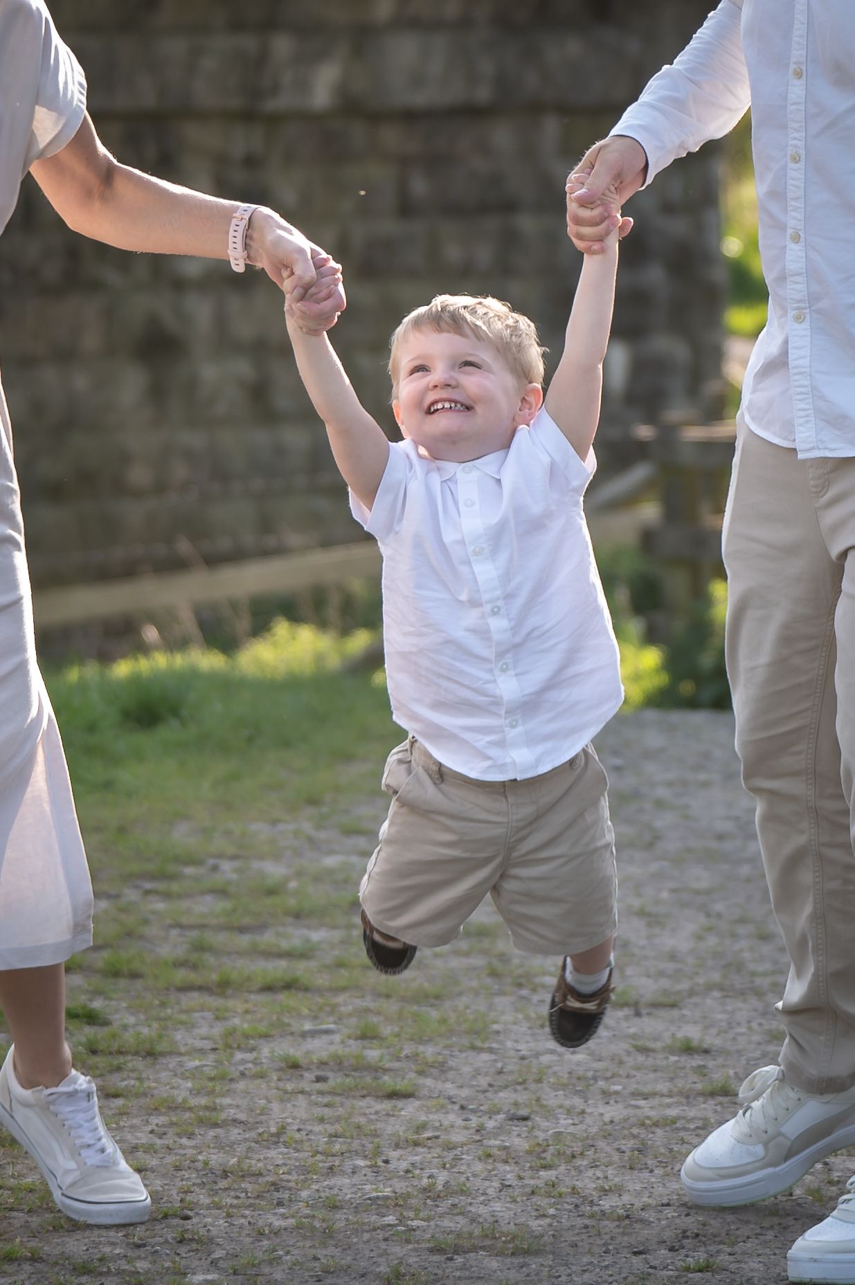 Young boy laughing as he is swung mid-air by both hands between two adults, wearing a white shirt and light trousers on a cobblestone path.