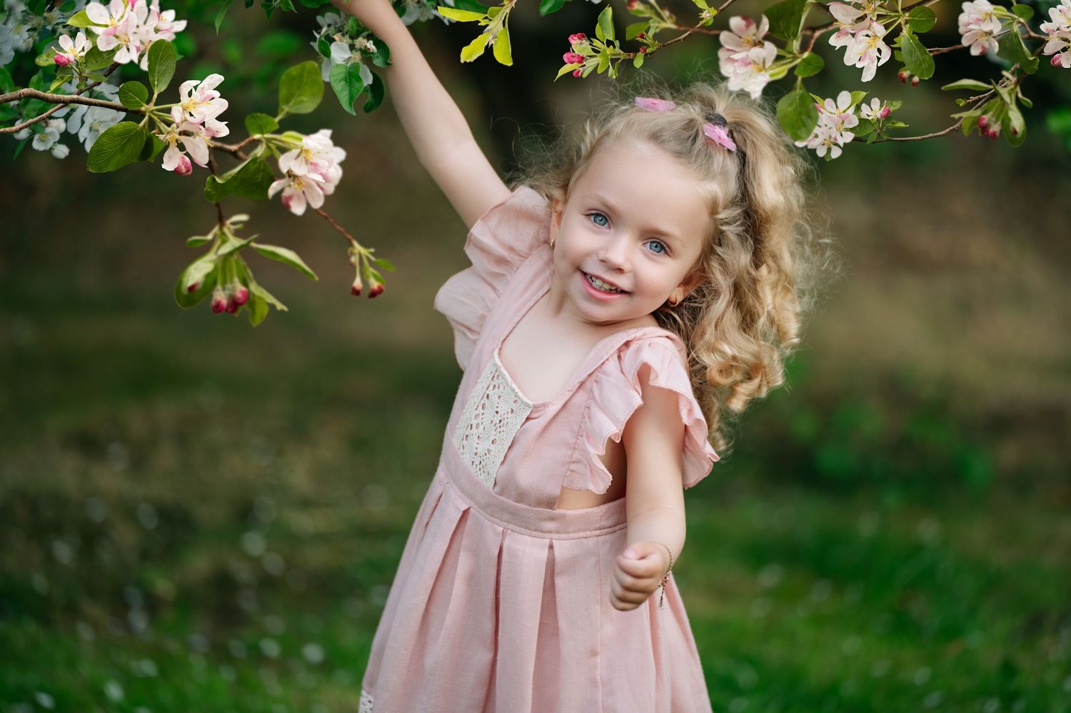 Joyful young girl reaching up toward blooming tree branches, smiling brightly in a pale pink dress among spring flowers.