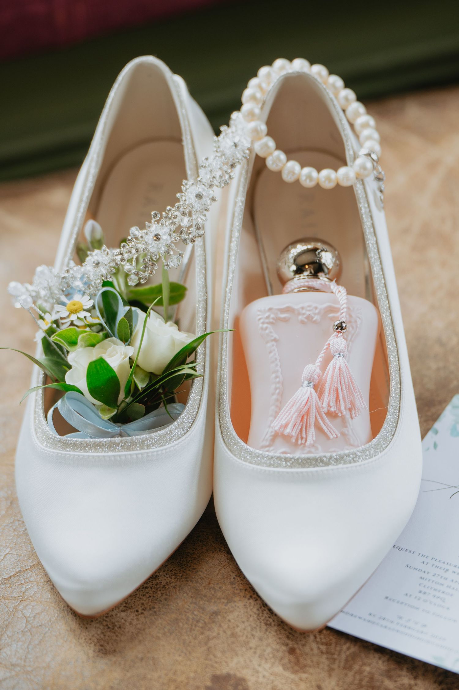 Close-up of white bridal shoes adorned with pearls, displayed with a delicate ring box and floral boutonnière on a wooden surface.