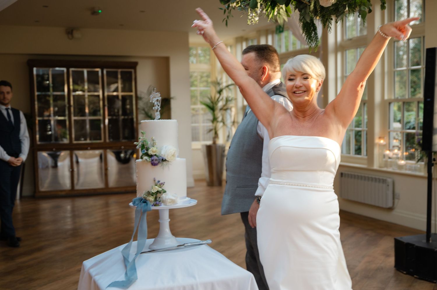 Bride joyfully raising her arms in celebration during the wedding reception, with the groom smiling beside her in a bright, elegant venue.