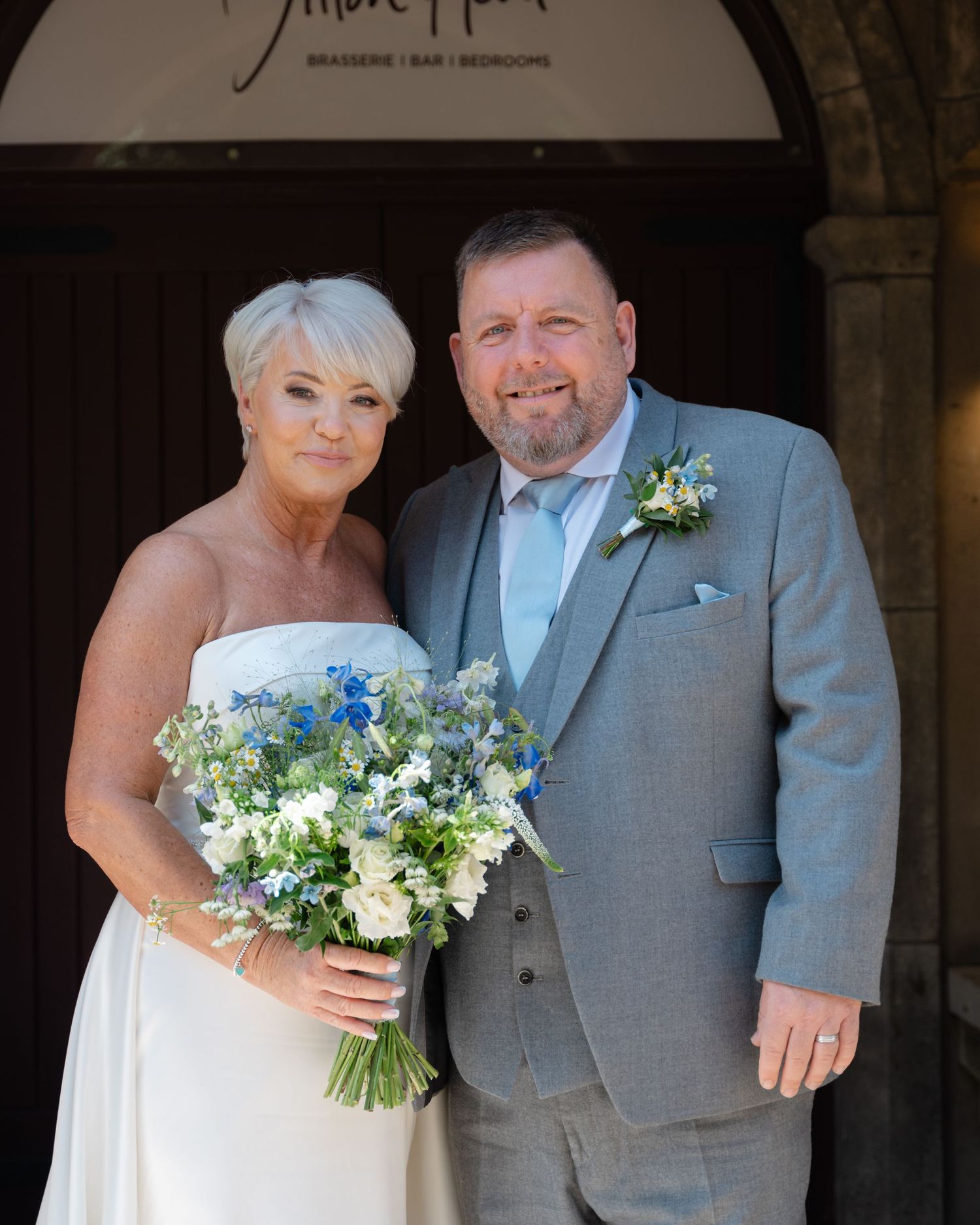 Bride and groom standing at the entrance of a venue, smiling and holding a bouquet of blue and white flowers, dressed in wedding attire.