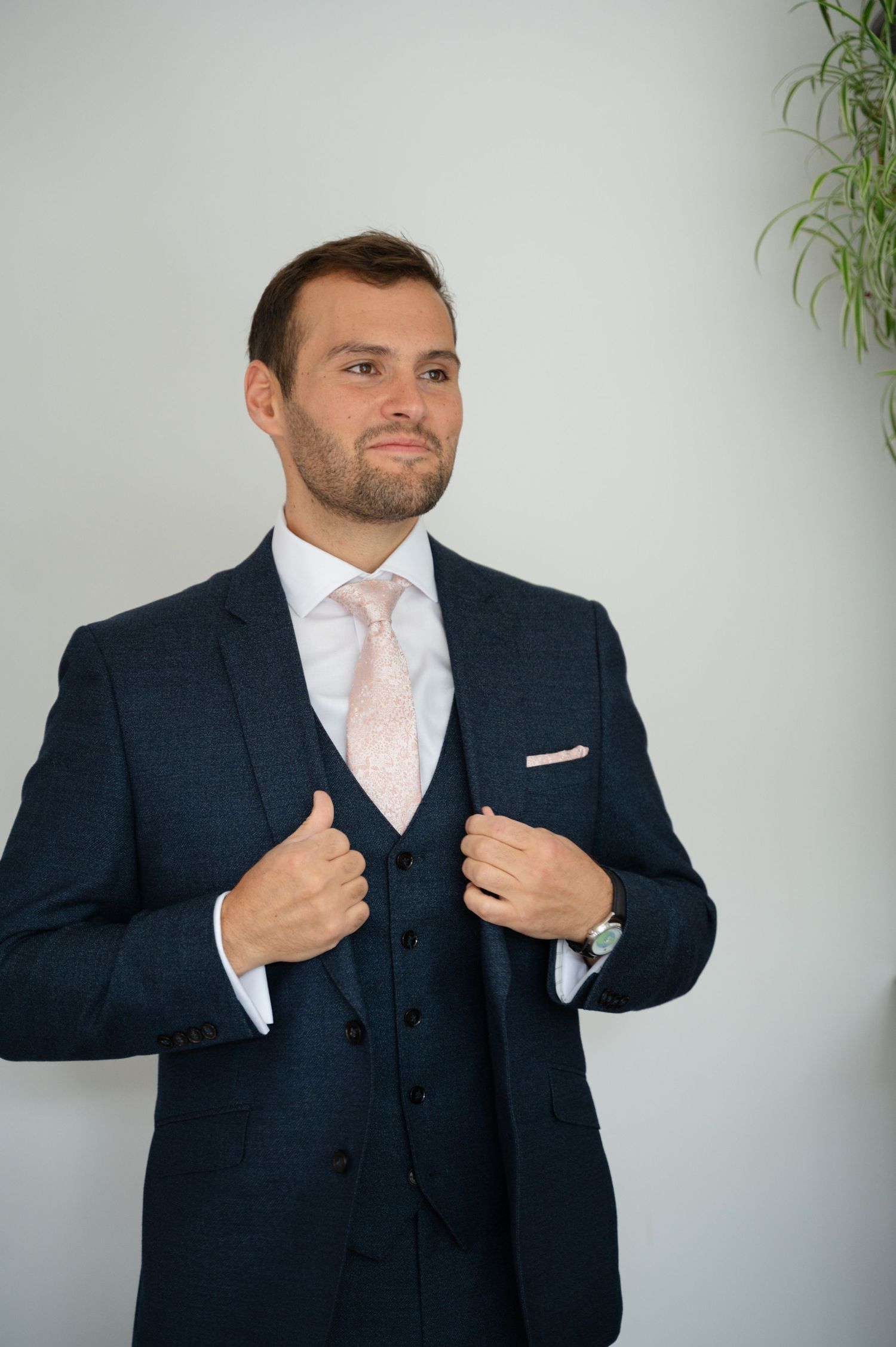 Groom buttoning up his suit jacket, standing against a white wall before the ceremony