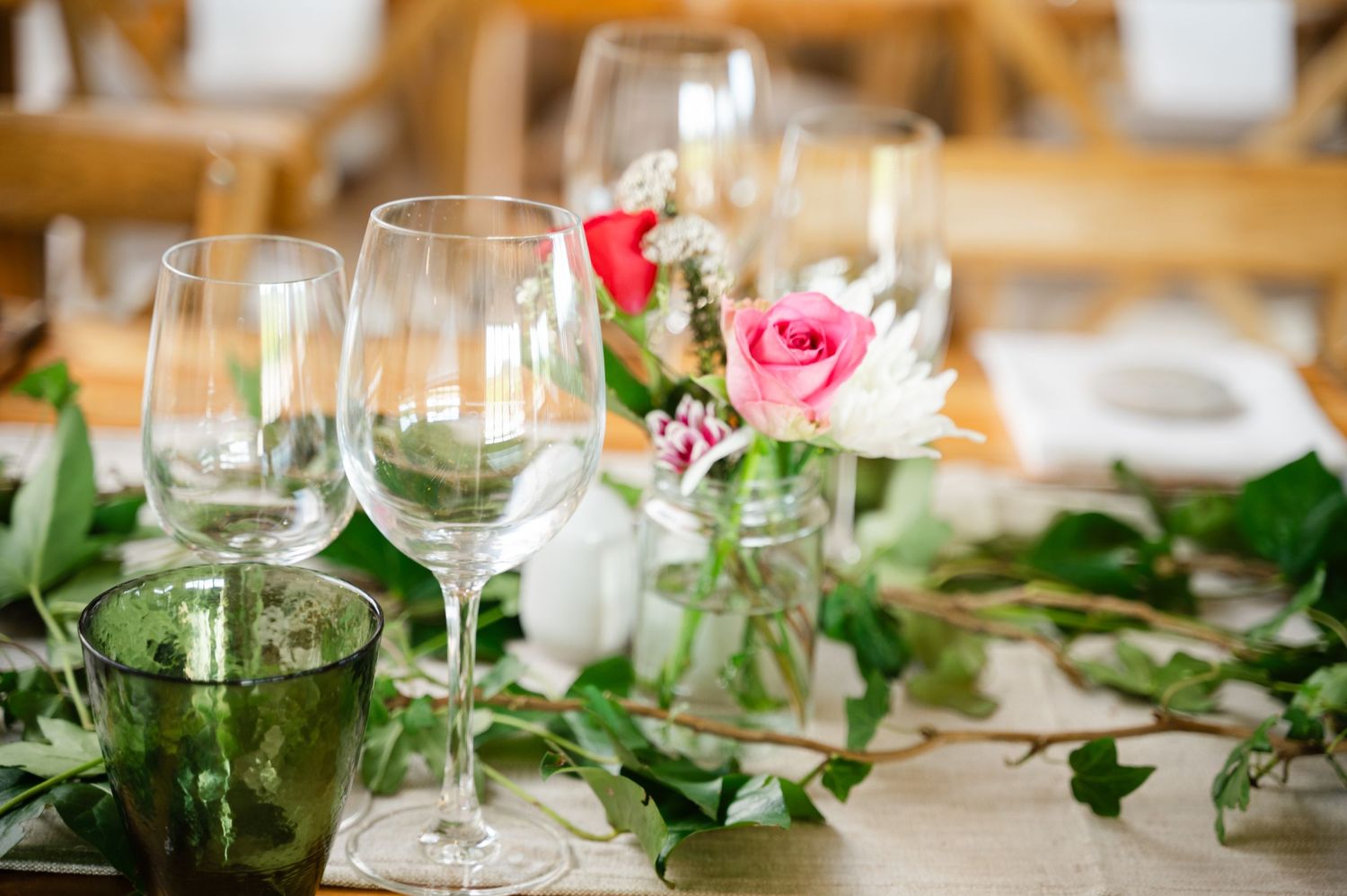 Close-up of wedding table setting with wine glasses, green foliage, and a single pink rose