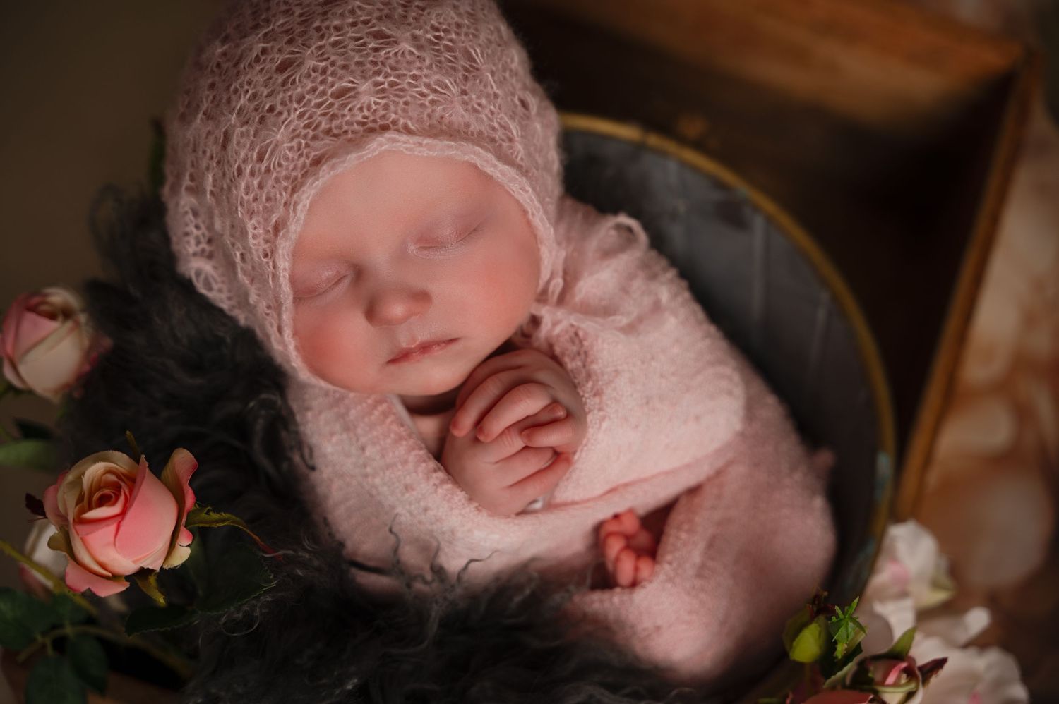 Newborn baby girl sleeping in a dark-toned basket, wrapped in a floral-patterned blanket with a lace bonnet, surrounded by soft shadows.