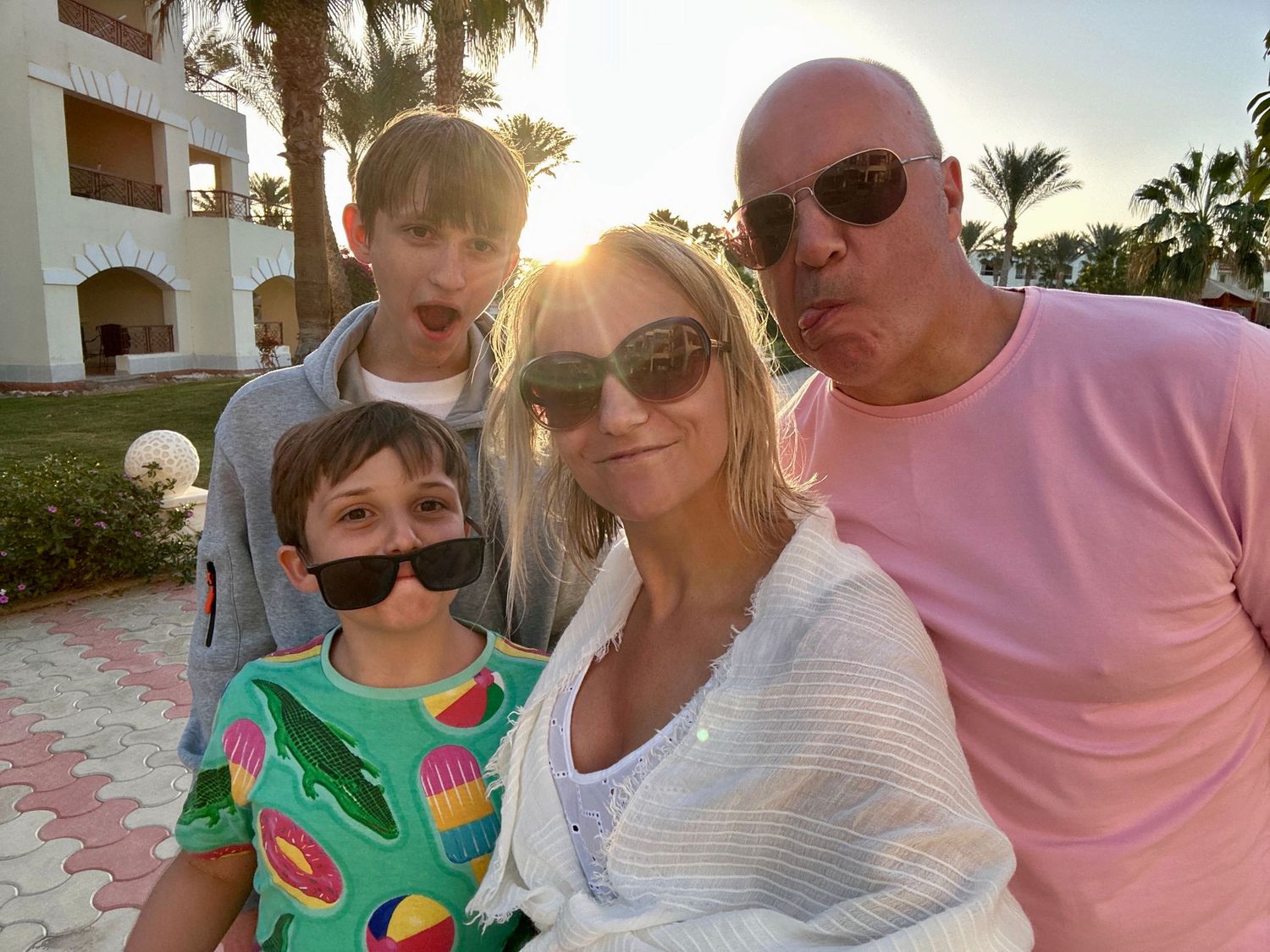 Family of three smiling outdoors on a sunny day, with palm trees in the background; parents and child dressed in colorful holiday clothing.
