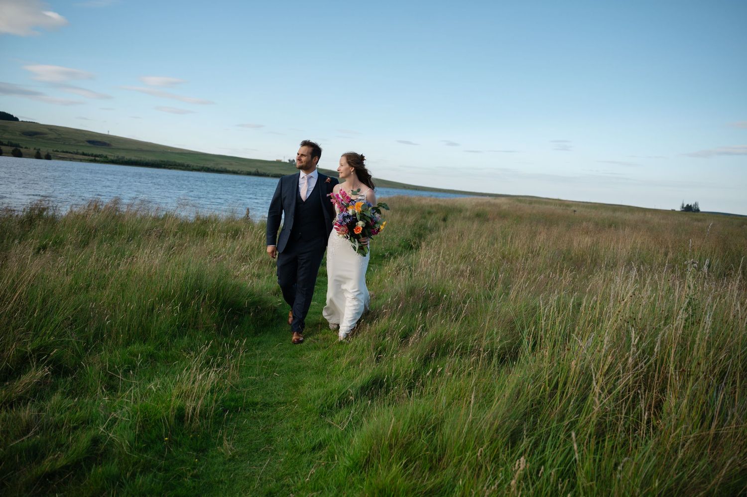Bride and groom walking through tall grass beside lake on wedding day