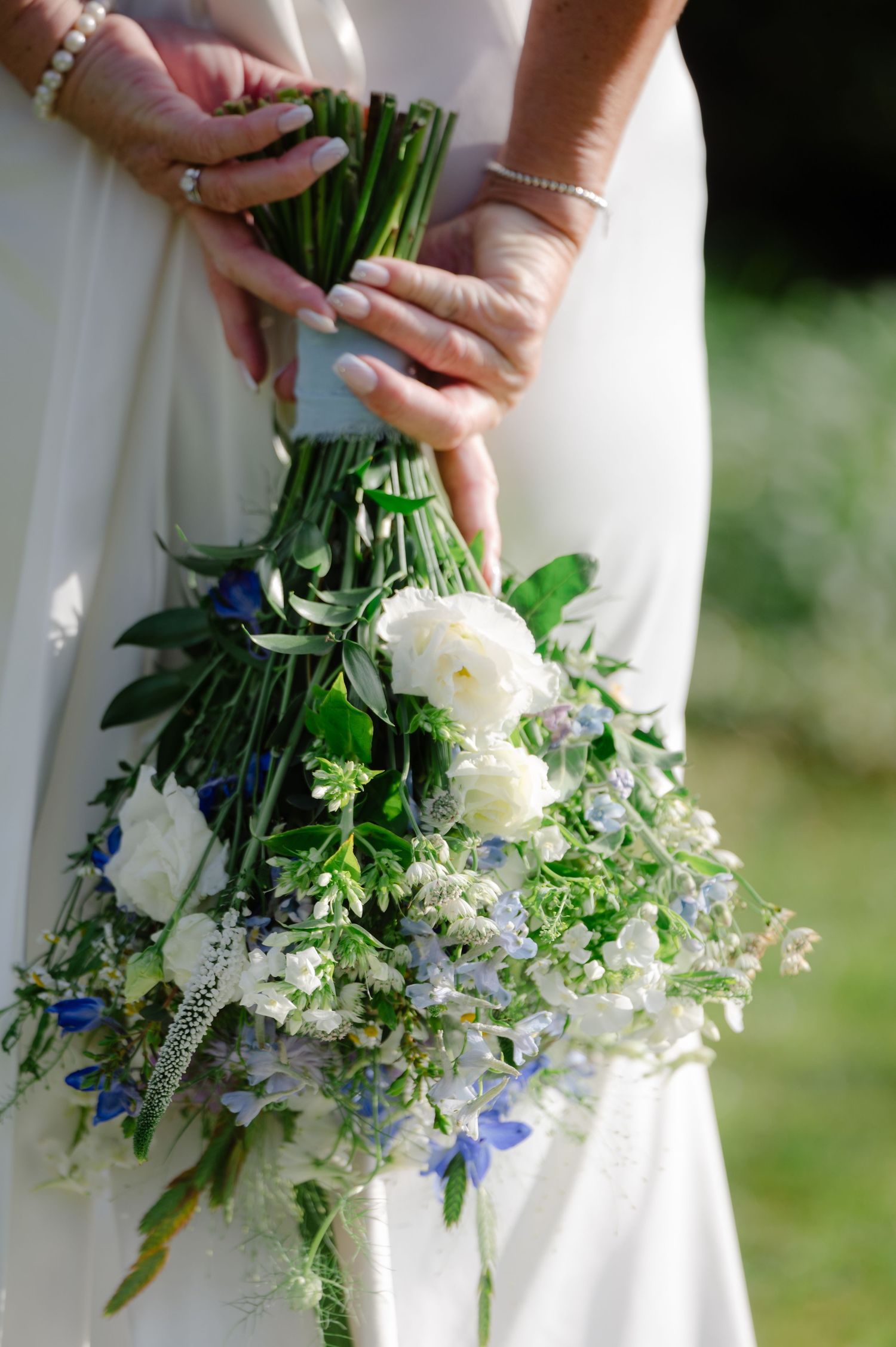 Close-up of bride holding a cascading bouquet of white and green flowers, with soft focus on the dress and outdoor background.