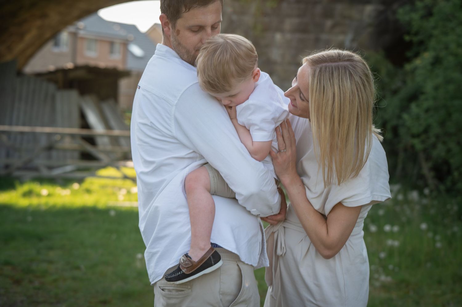 Mother, father, and young child standing together in a garden, smiling and cuddling, with sunlight filtering through behind them.