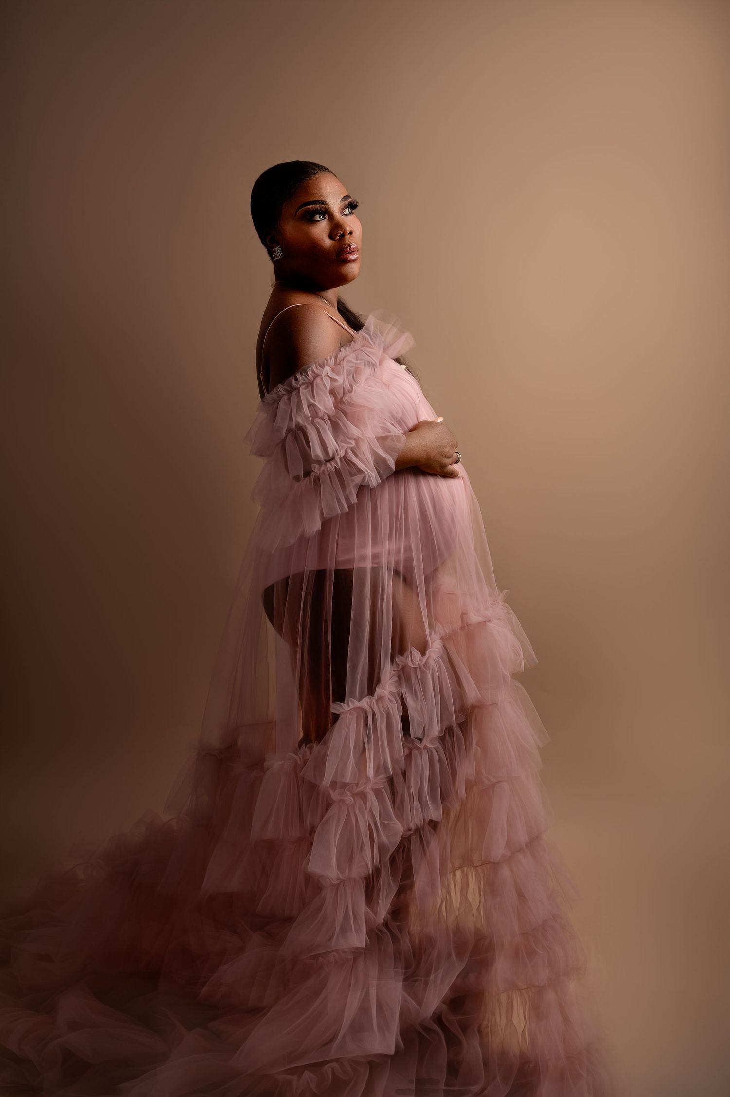 Woman in a dramatic pink tulle gown standing in profile against a beige backdrop, softly lit to create a moody, elegant atmosphere.