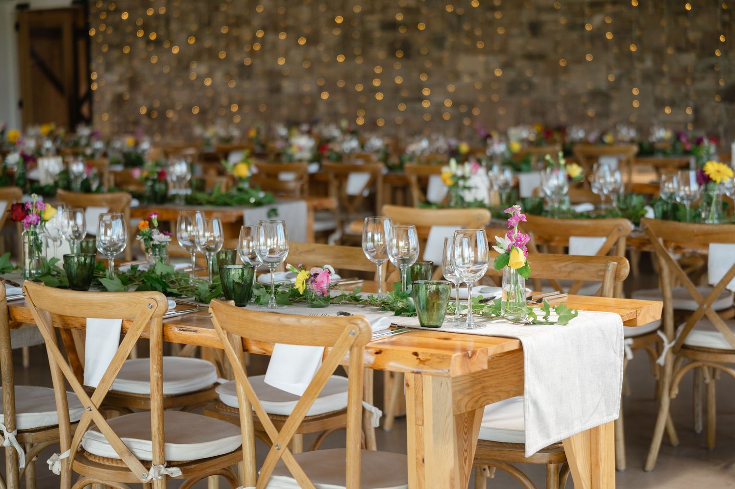 Long wooden tables set up for a wedding reception with greenery and candles in a barn venue