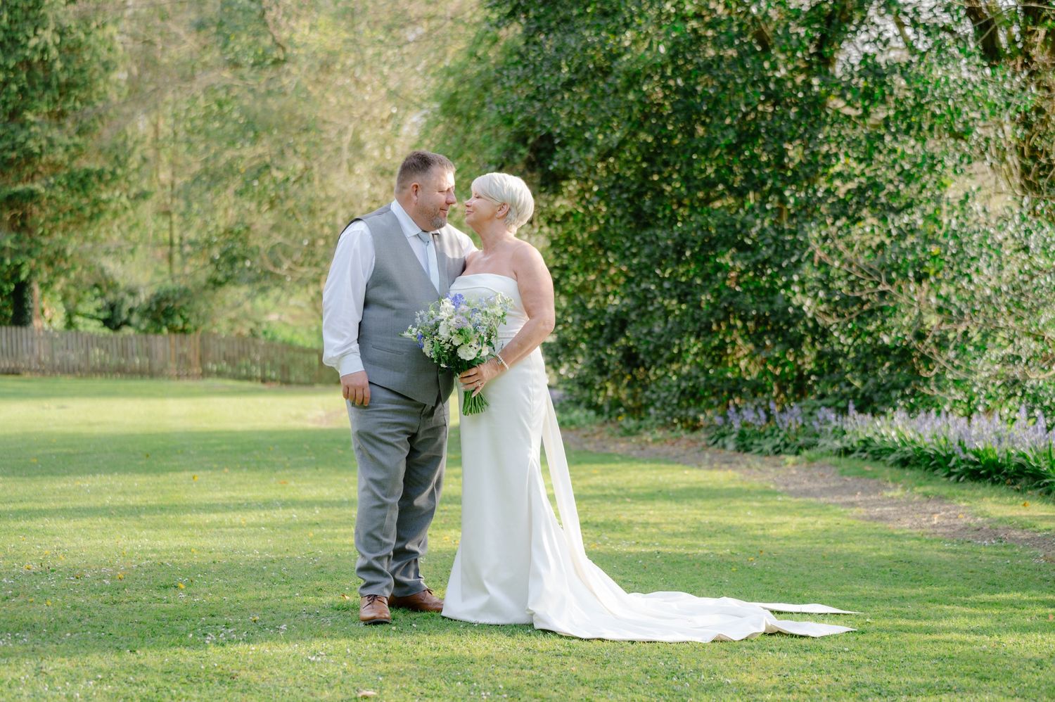 Bride smiling up at the groom while standing close together in a scenic countryside setting, with bouquet in hand and warm sunset tones.
